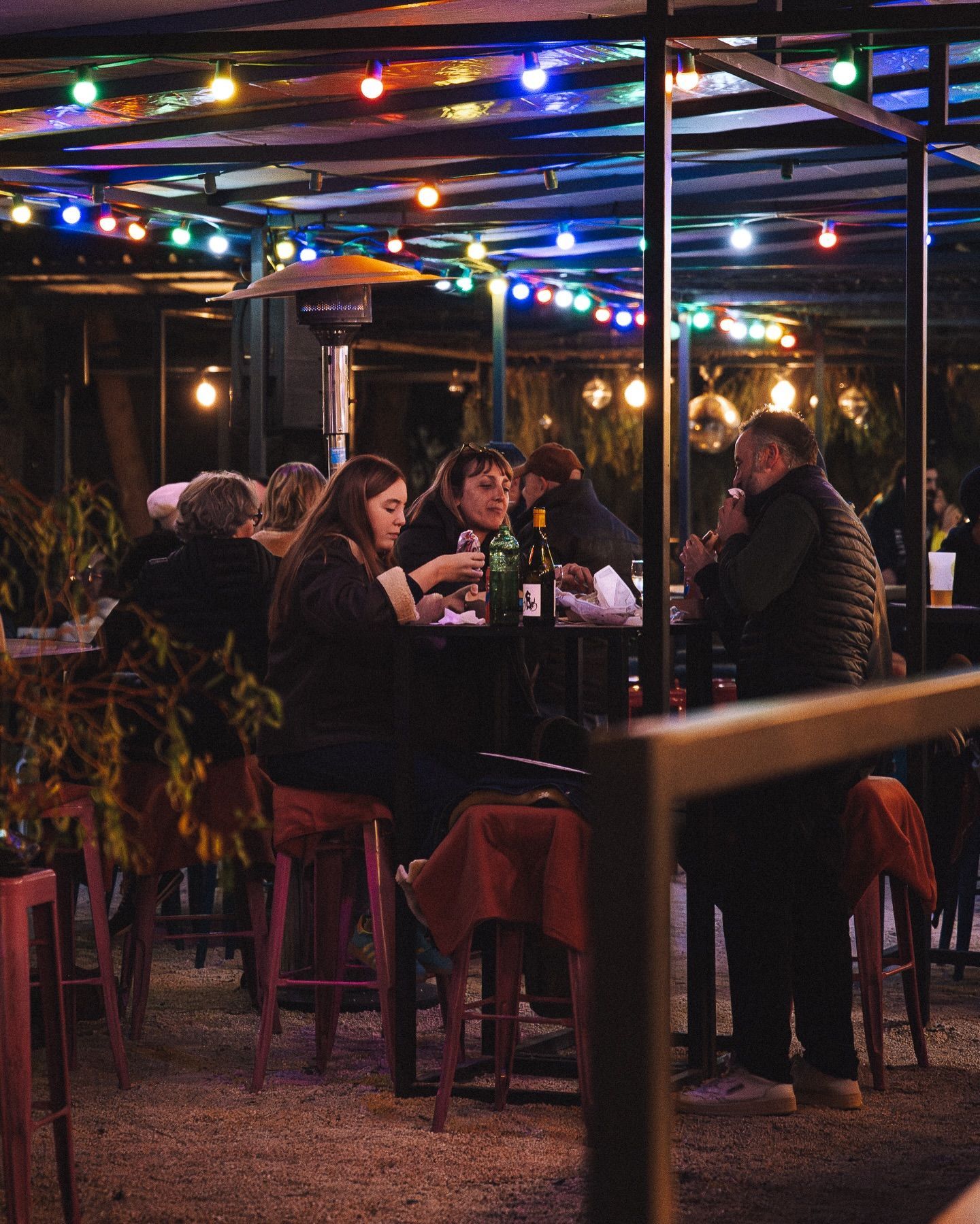 Des clients sont assis à des tables hautes dans un restaurant en plein air illuminé par des guirlandes lumineuses