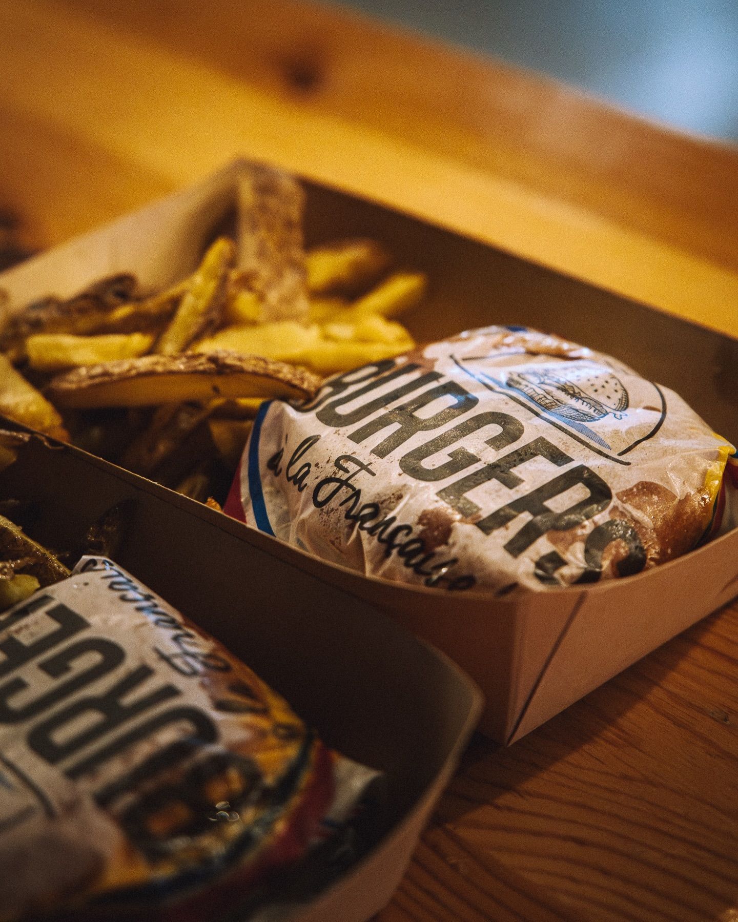 Deux hamburgers emballés dans du papier et une portion de quartiers de pommes de terre servis dans des barquettes en carton sur une table en bois