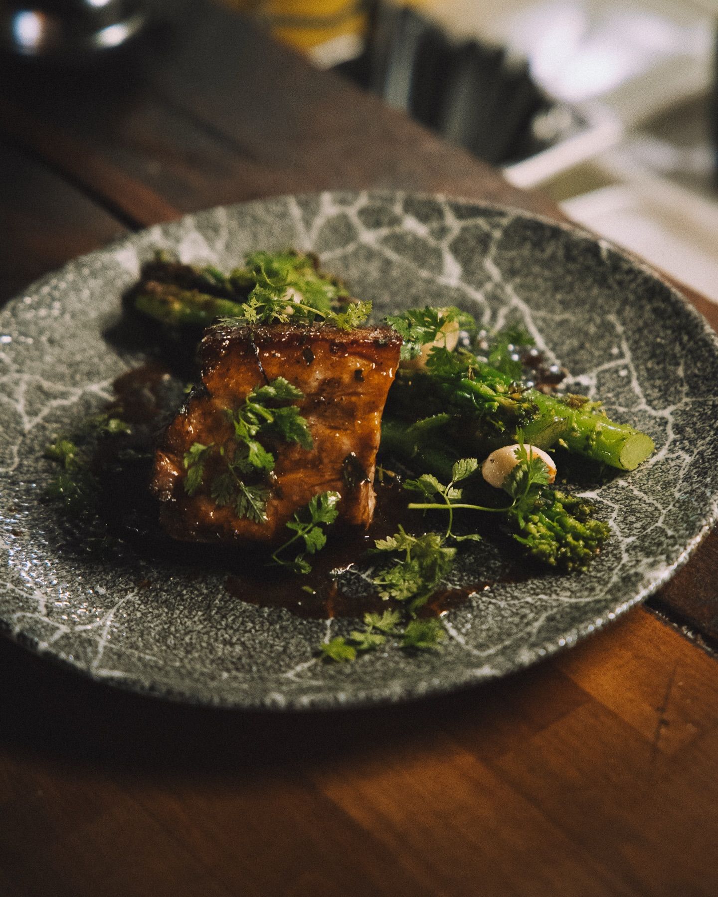 Un morceau de viande rectangulaire glacé, servi avec des asperges rôties sur une assiette texturée grise à motif craquelé