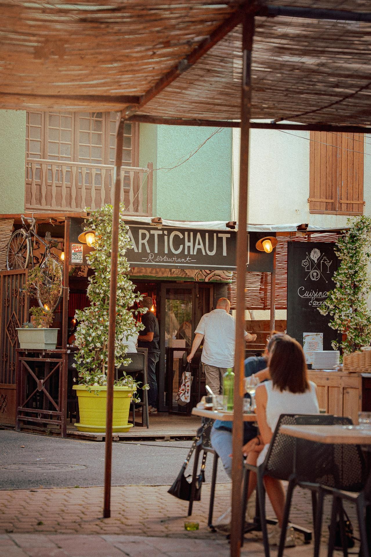 Restaurant avec des places assises en extérieur, une jardinière jaune et des clients dînant sous une terrasse ombragée