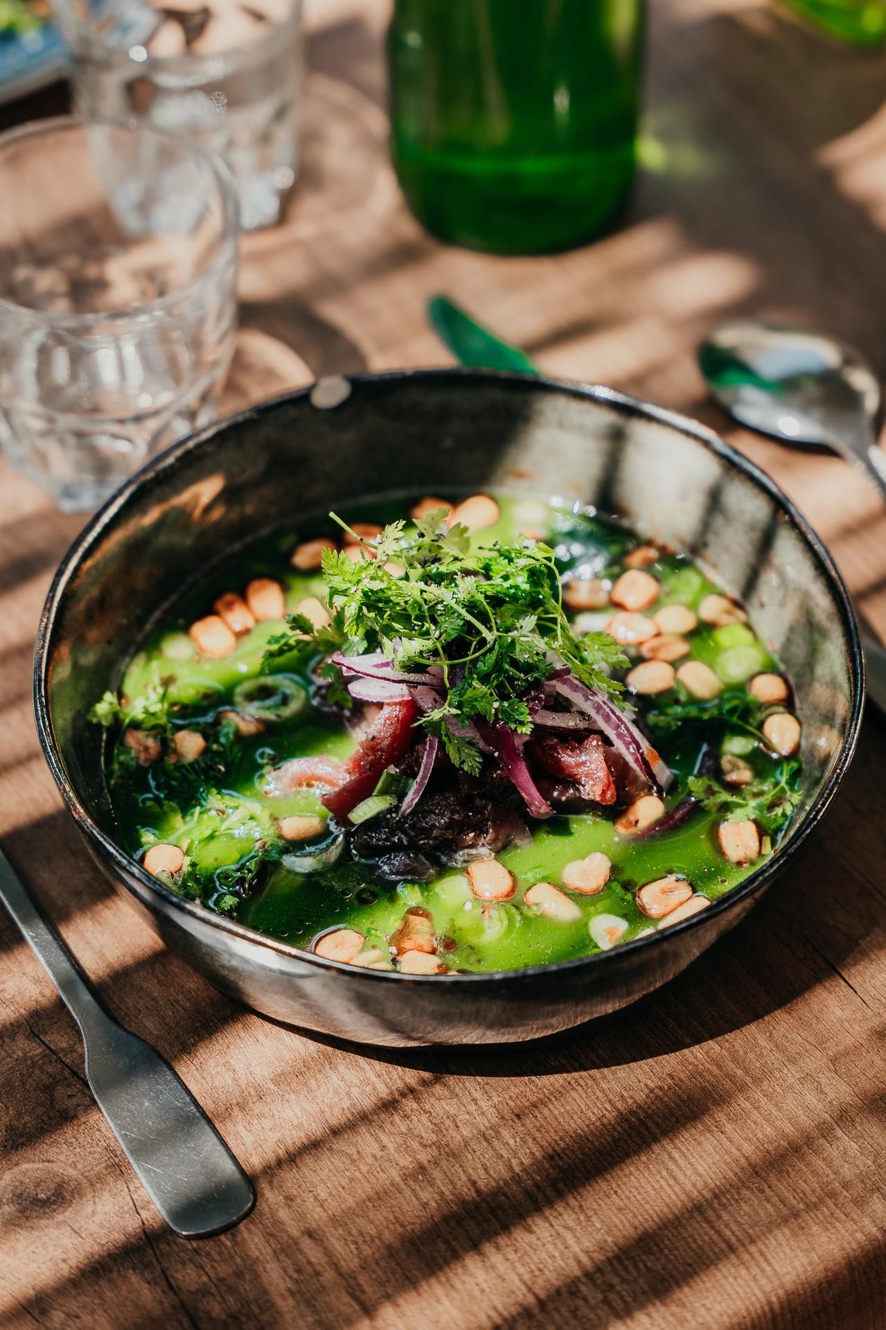 Un bol de soupe verte garni d'herbes, d'oignon rouge et de croûtons, posé sur une table en bois avec des verres et des couverts