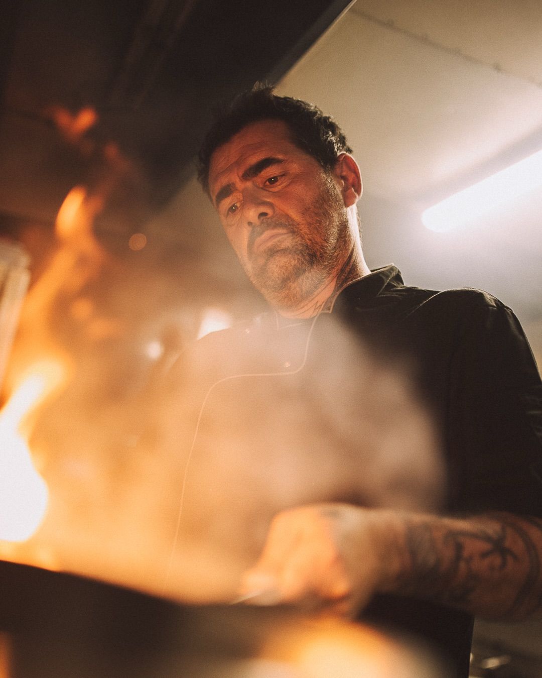 Un chef en chemise noire cuisine sur un feu de bois dans une cuisine, la vapeur s'échappant et le feu étant visible au premier plan.
