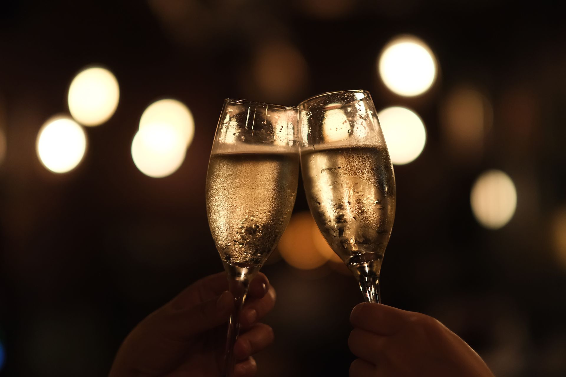Deux mains lèvent des flûtes de champagne, sur un fond sombre illuminé de lumières floues
