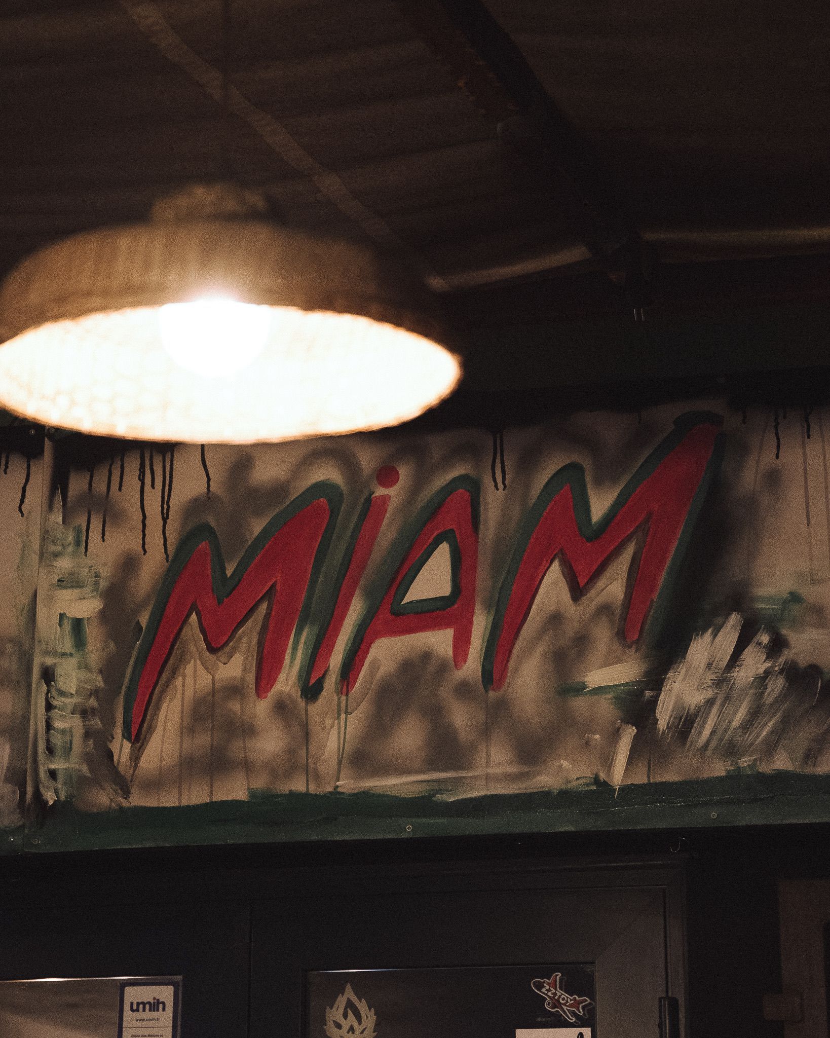 Le mot MIAM est peint à la bombe en lettres rouges avec un contour noir sur un mur sombre et texturé, sous une suspension lumineuse