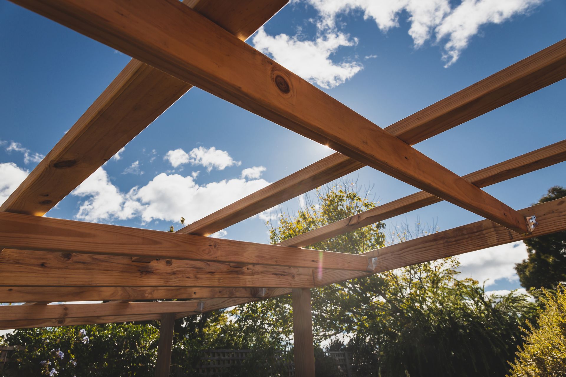 Des poutres de pergola en bois encadrent un ciel d'un bleu éclatant, avec des nuages ​​et des arbres à l'horizon.