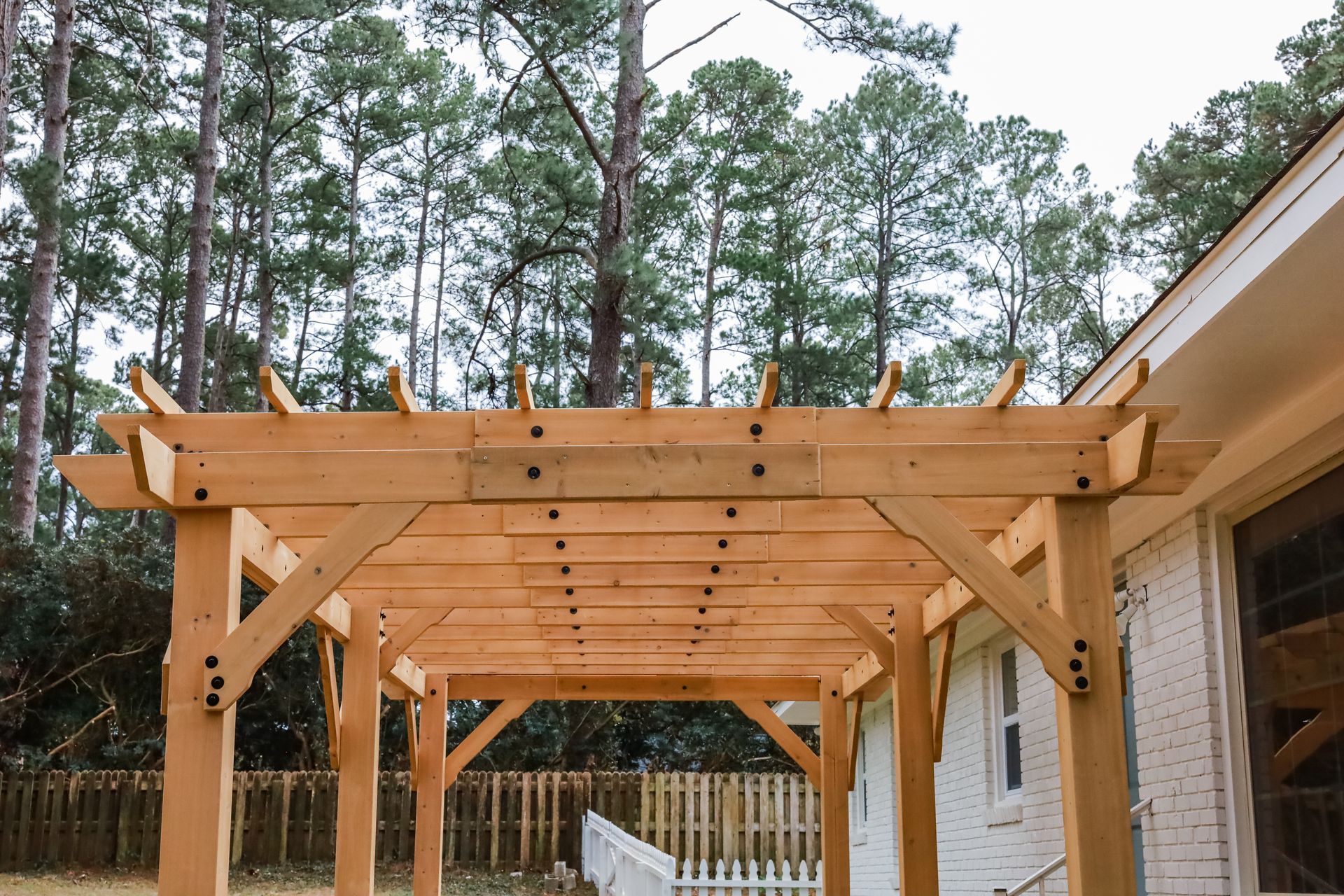 Pergola en bois en construction à côté d'une maison, avec des pins en arrière-plan.