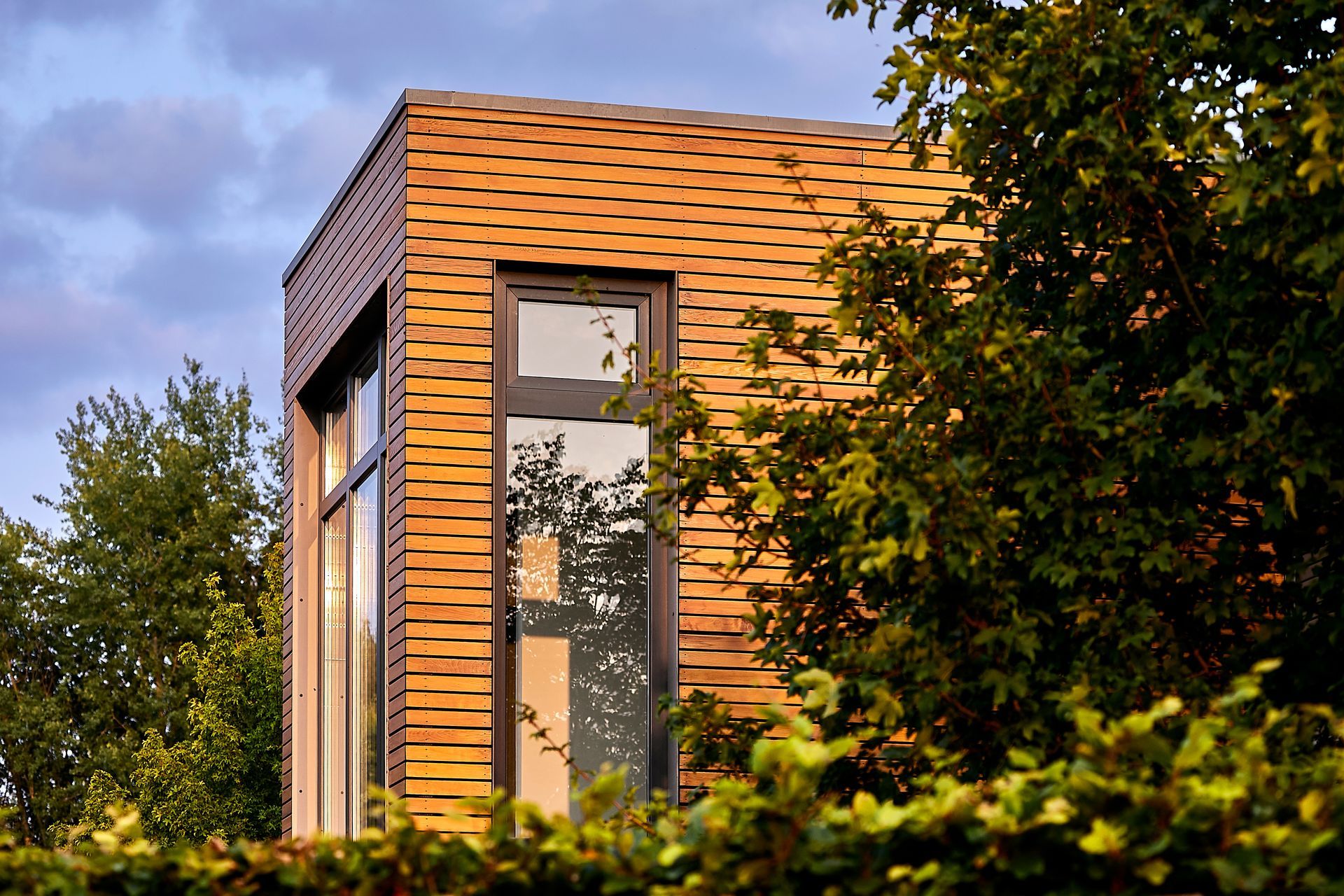 Façade moderne en bois d'une maison avec de hautes fenêtres, encadrée par des arbres et de la verdure au coucher du soleil