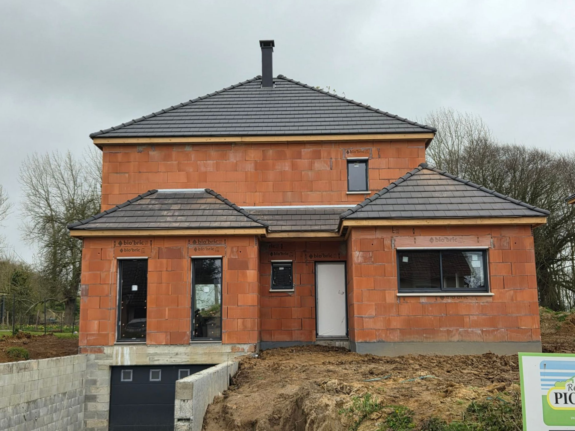 Maison en briques inachevée avec toit gris, porche et chantier devant.