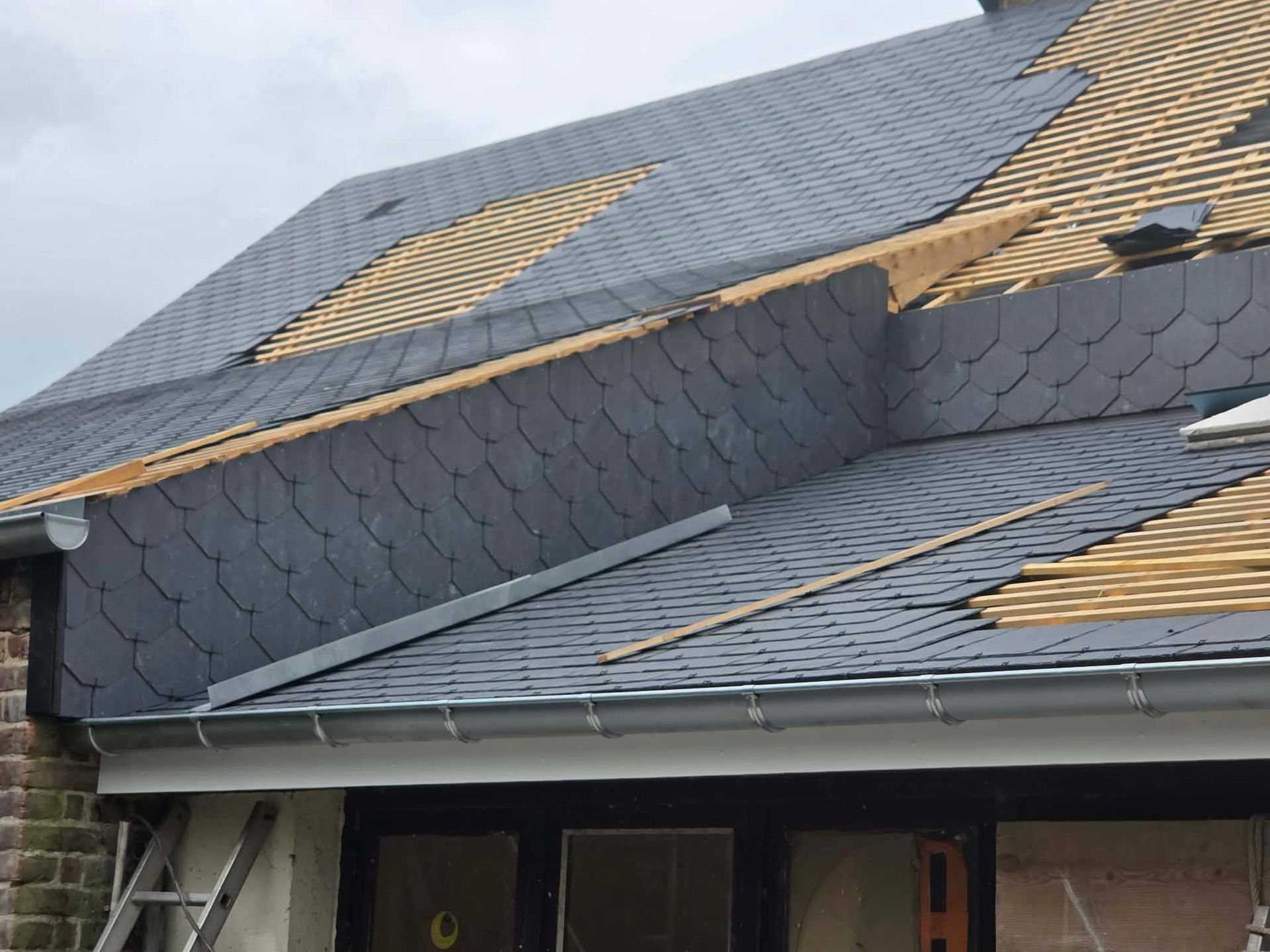 Toiture gris foncé endommagée, avec des tuiles manquantes et une sous-couche exposée au-dessus de l'entrée de la maison