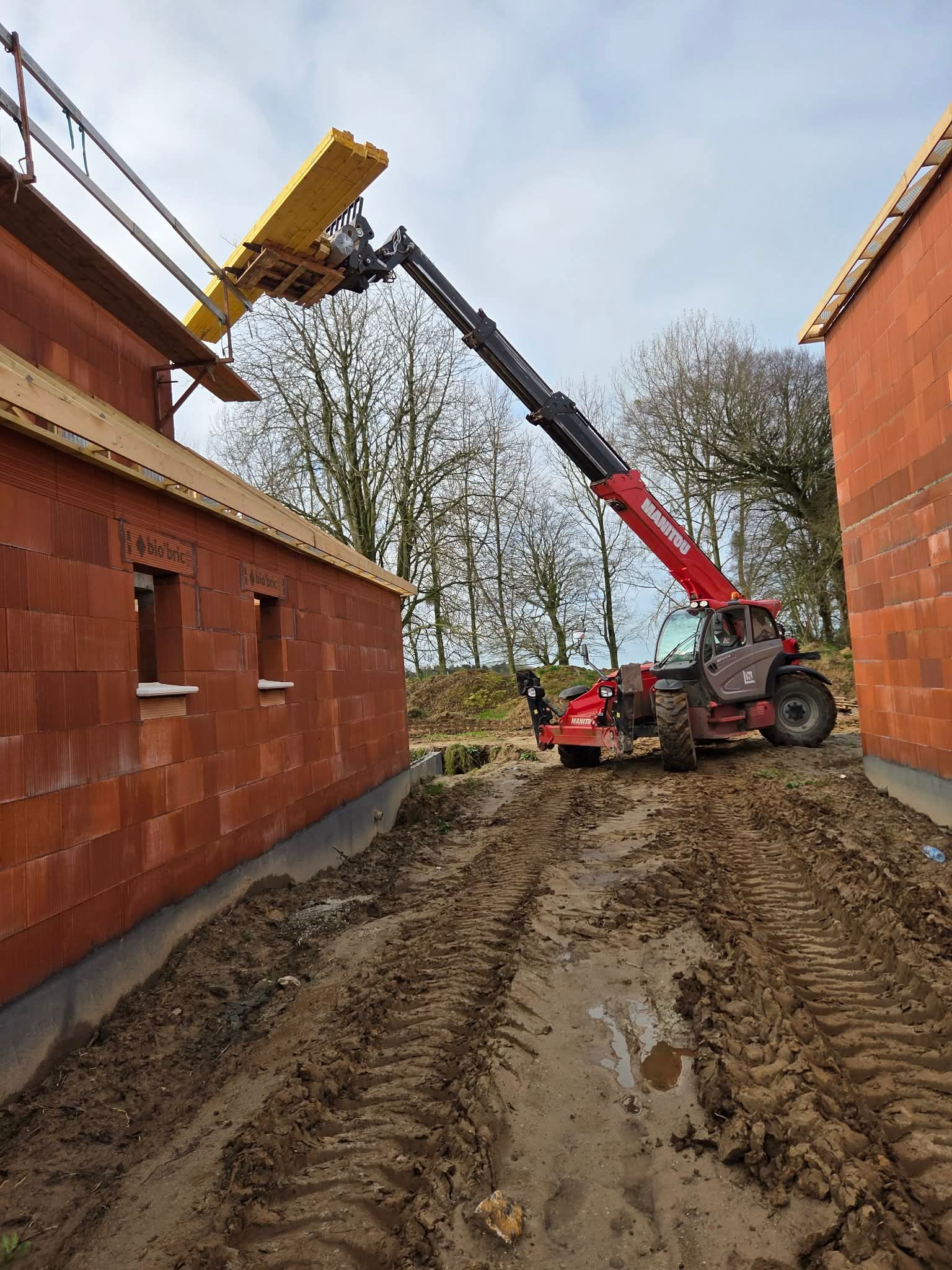 Un chariot télescopique rouge soulève une ferme de toit jaune à côté d'un bâtiment en briques sur un chantier boueux.