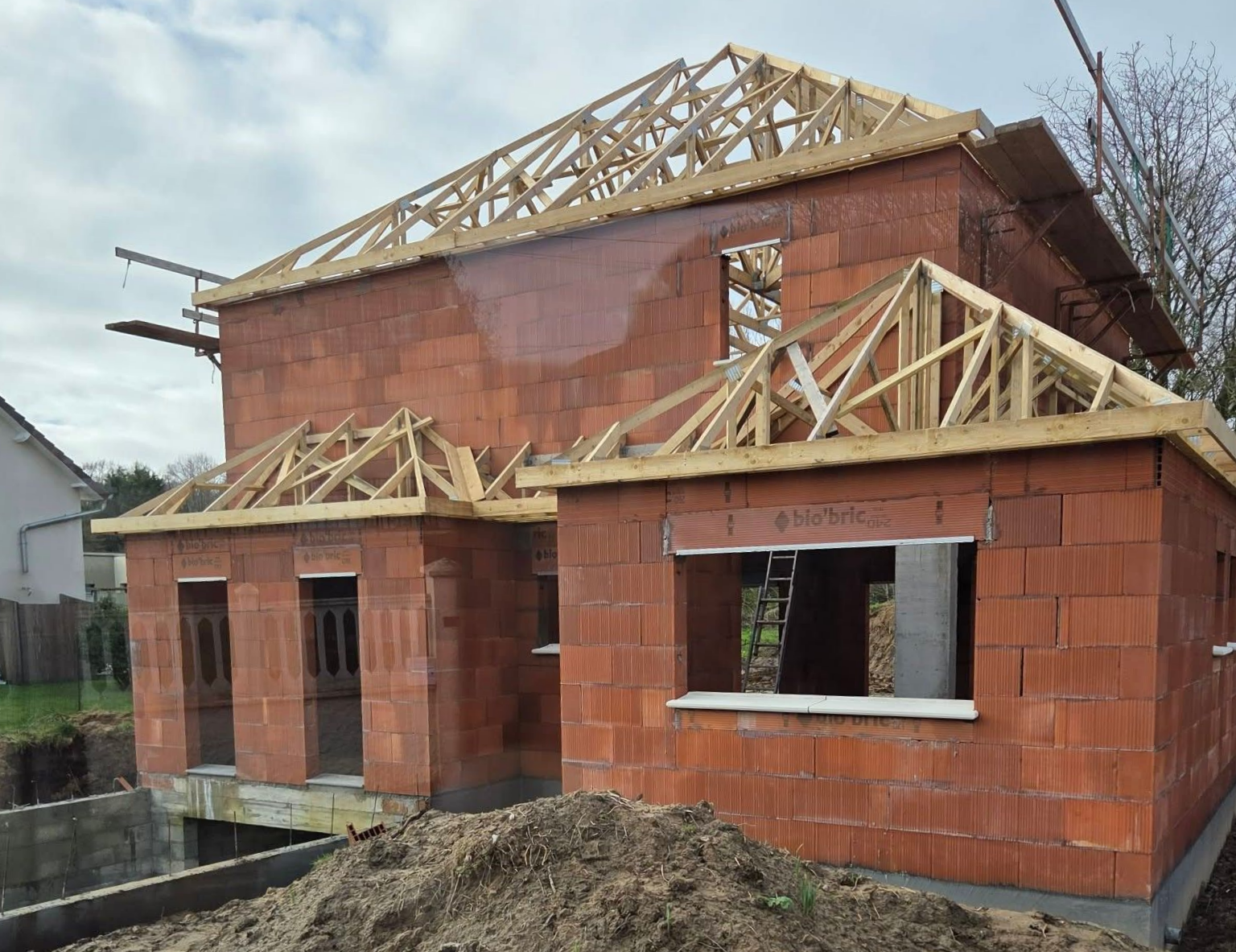 Maison en briques rouges partiellement construite, avec charpente apparente et murs inachevés, en cours de construction.