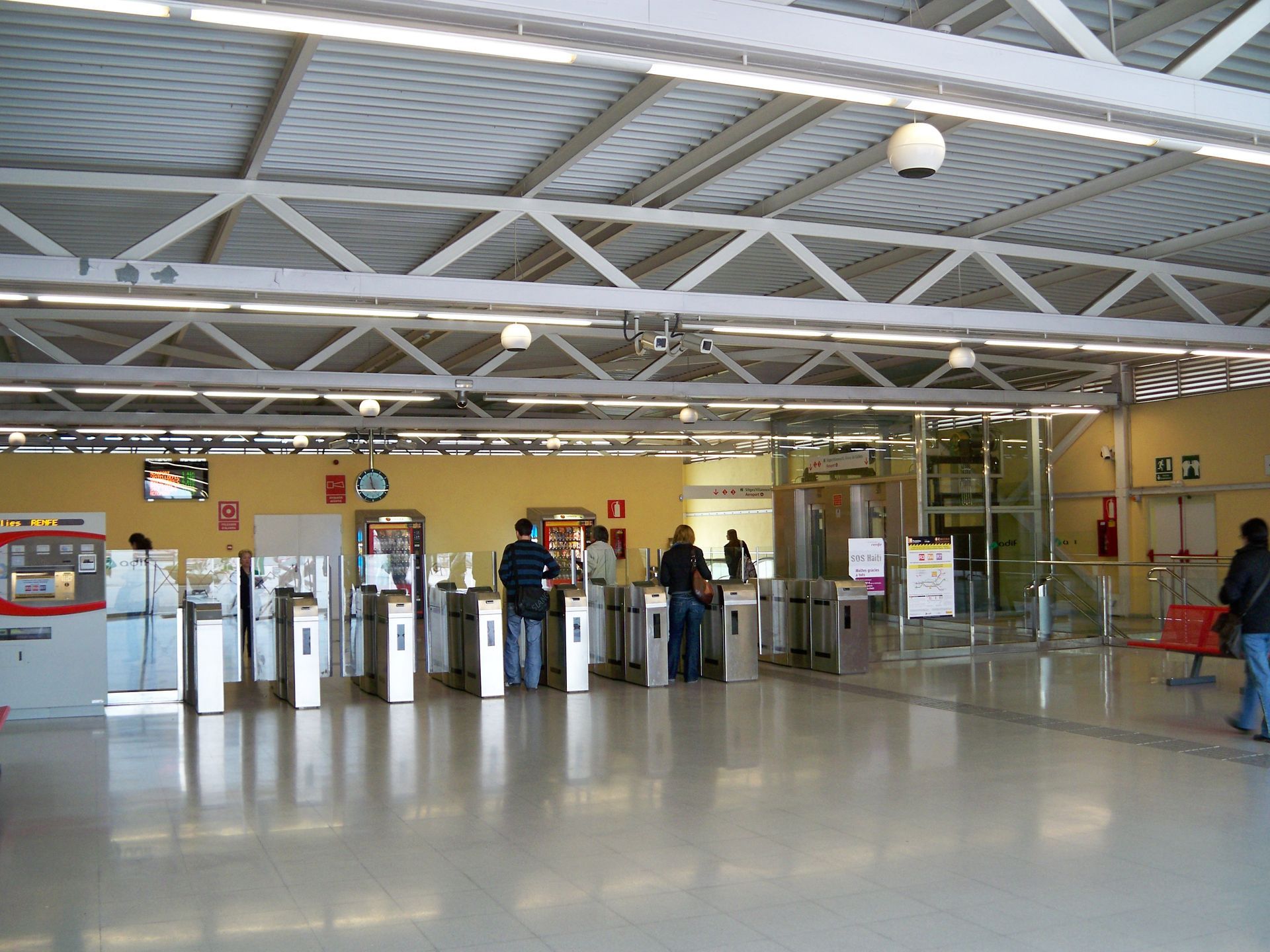 Personas en una estación de tránsito con torniquetes. Interior bien iluminado con techo metálico, máquinas expendedoras de billetes y ascensores de cristal.