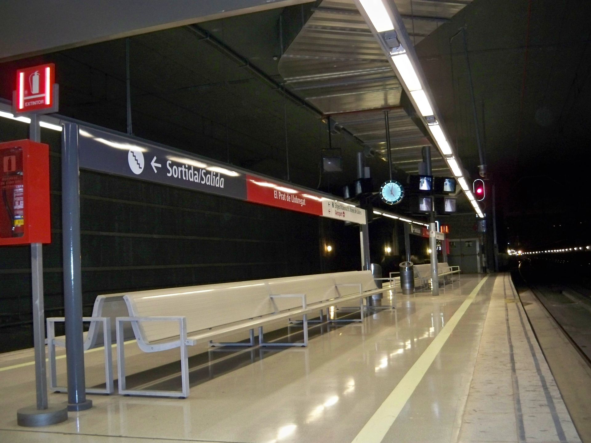 Andén del metro de noche, con un banco largo, señalización y una caja de alarma contra incendios de color rojo.