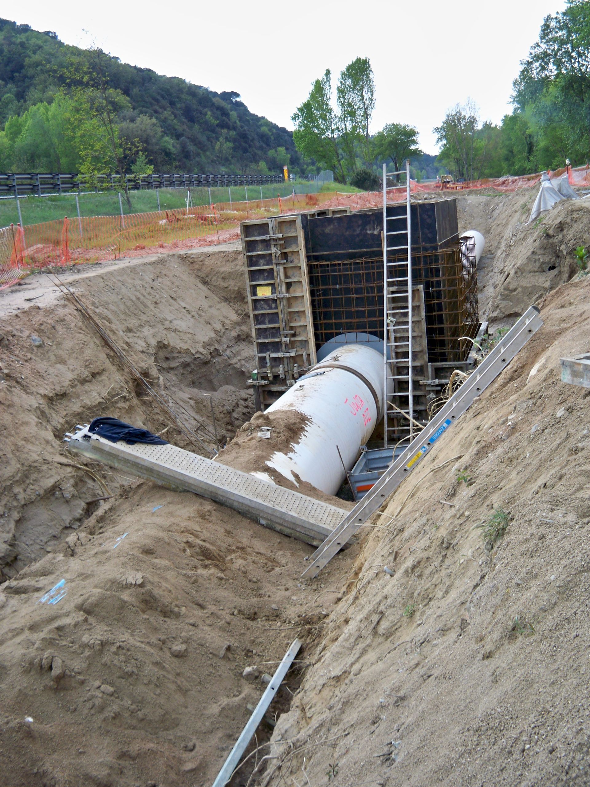 Sitio de construcción con tubería grande en zanja; muro de contención en construcción, escalera y cerca de seguridad presentes.