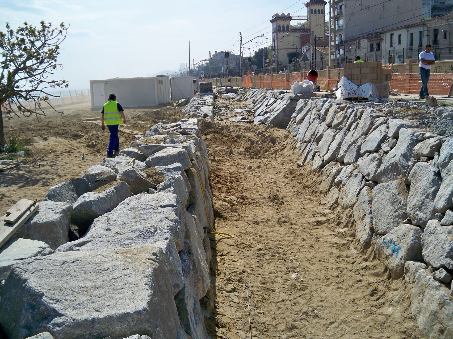 Sitio de construcción a lo largo de una playa de arena; muros de piedra y trabajadores; edificios al fondo.