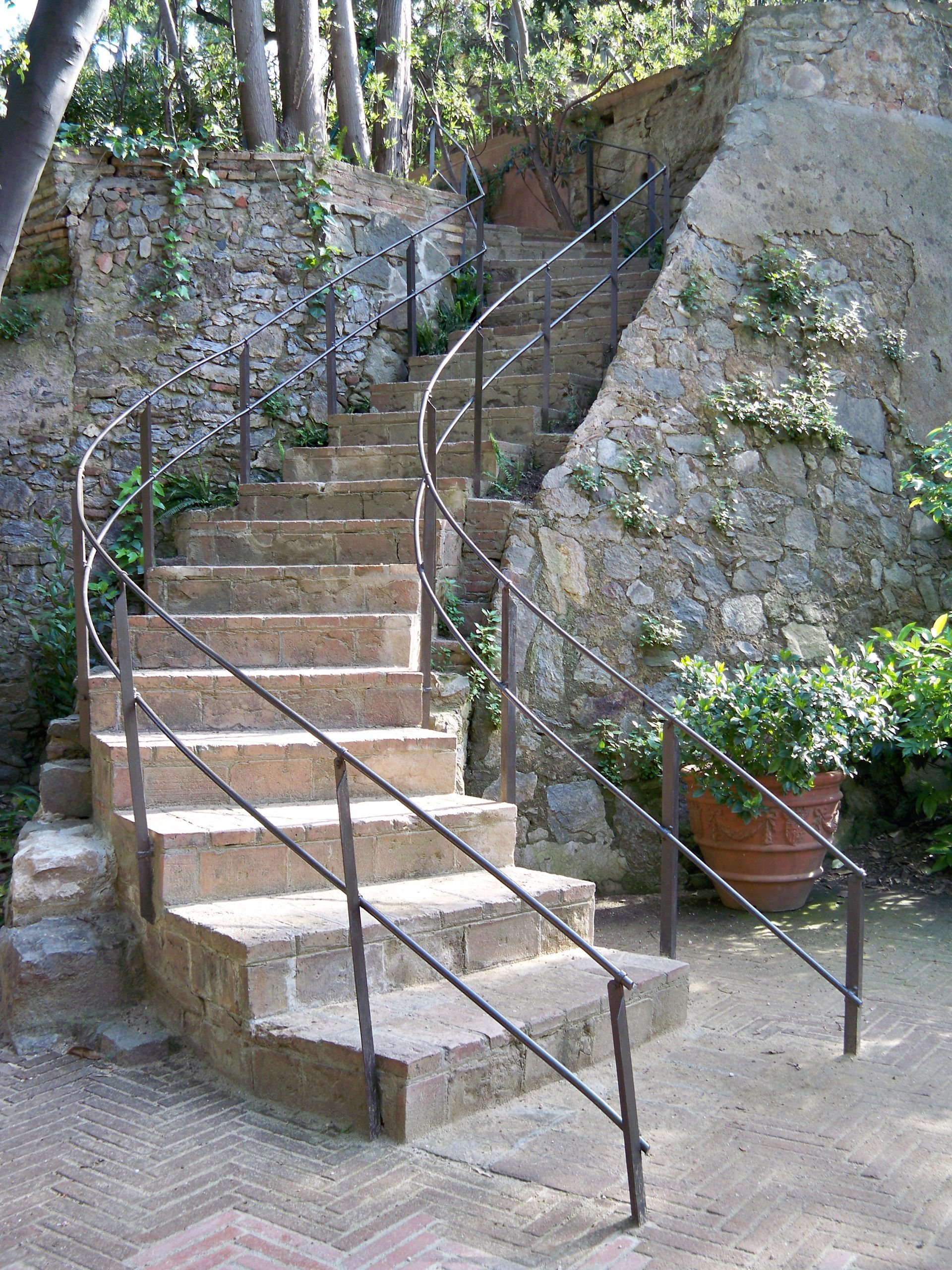 Una escalera de piedra con barandilla de hierro asciende por un entorno de jardín, con paredes de piedra y plantas en macetas.