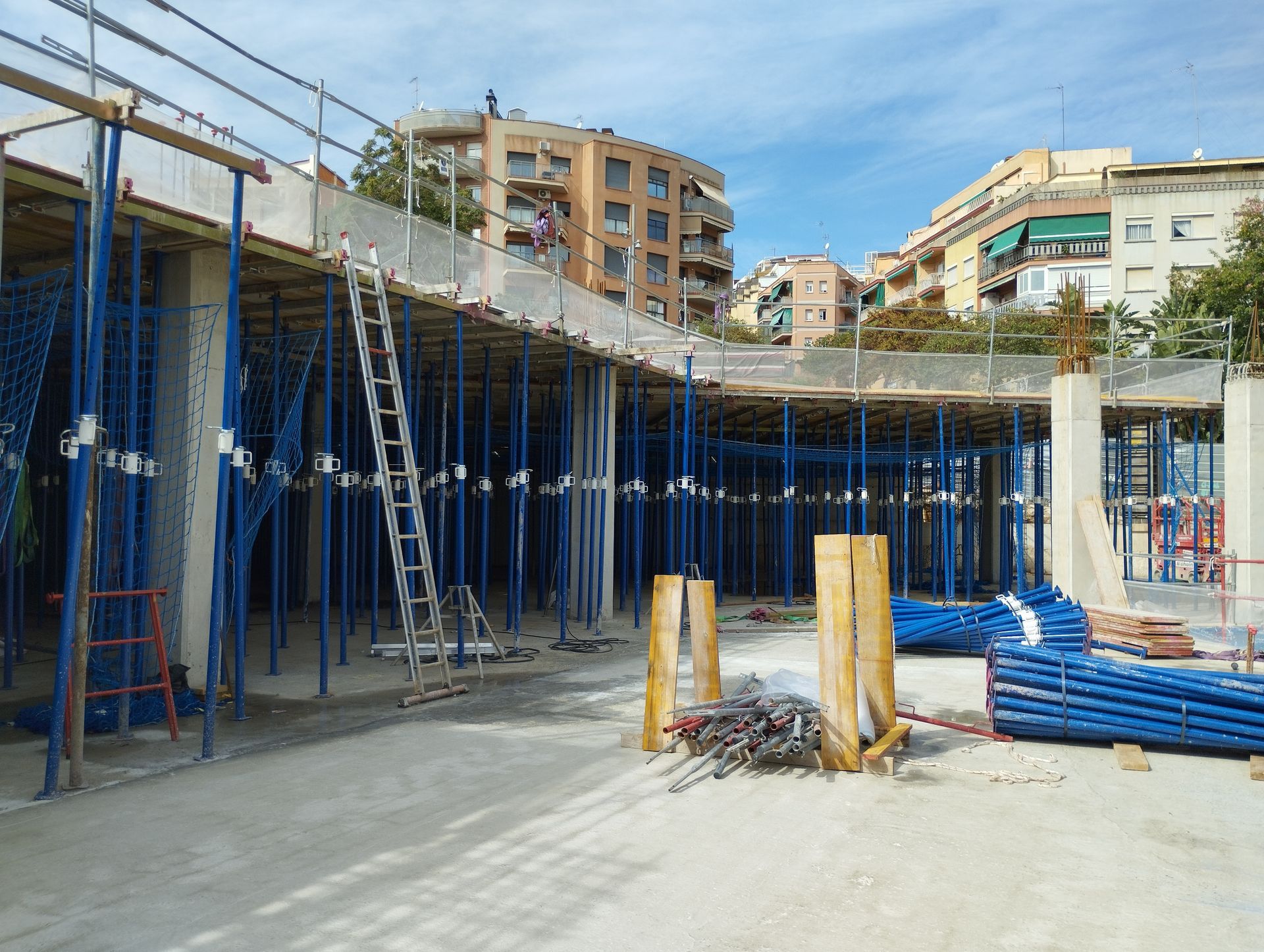 Sitio de construcción con andamios azules que sostienen una estructura de hormigón; edificios residenciales en el fondo.