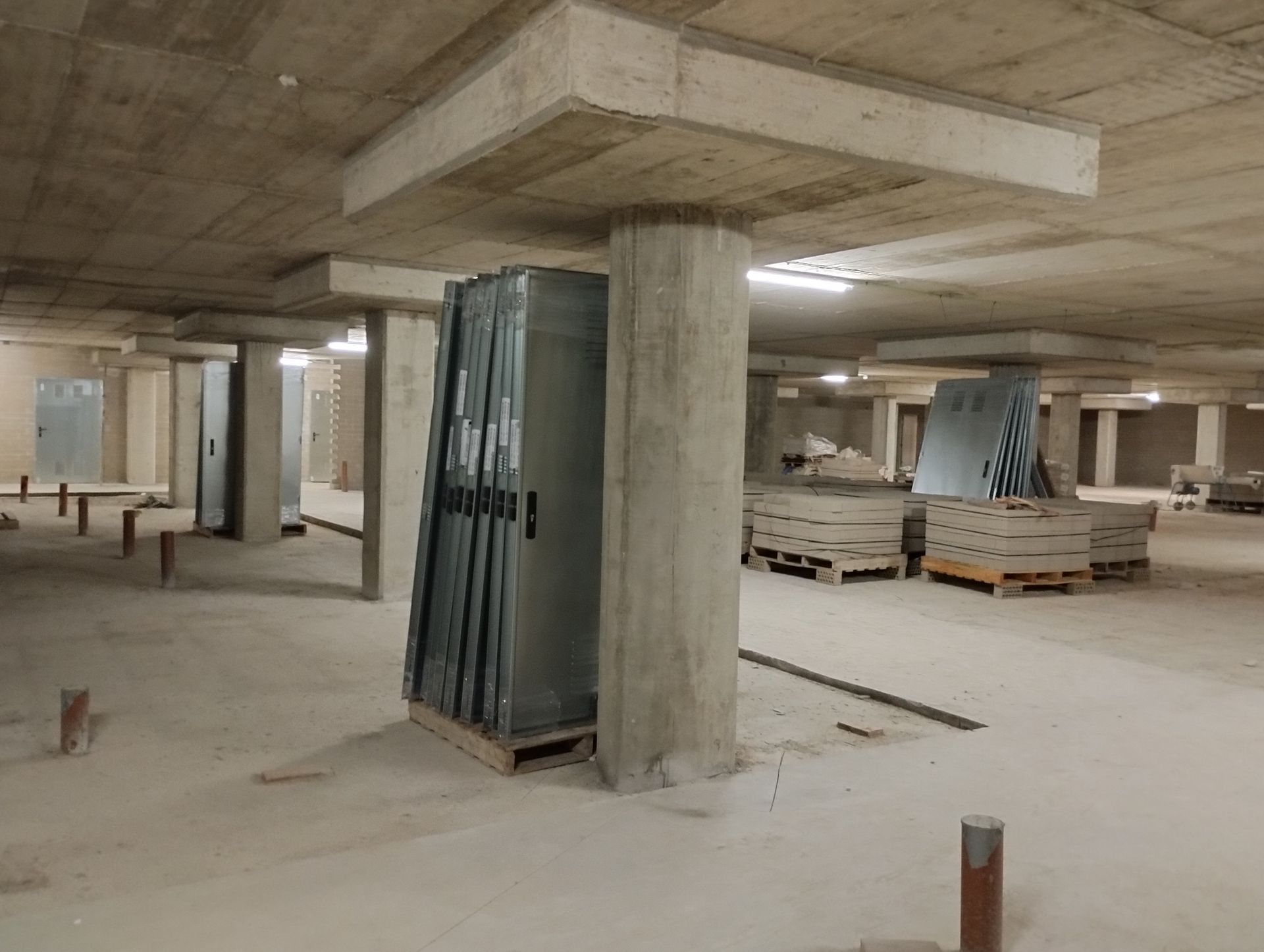 Obra de construcción de aparcamiento de hormigón con puertas apiladas sobre palets y columnas de soporte.