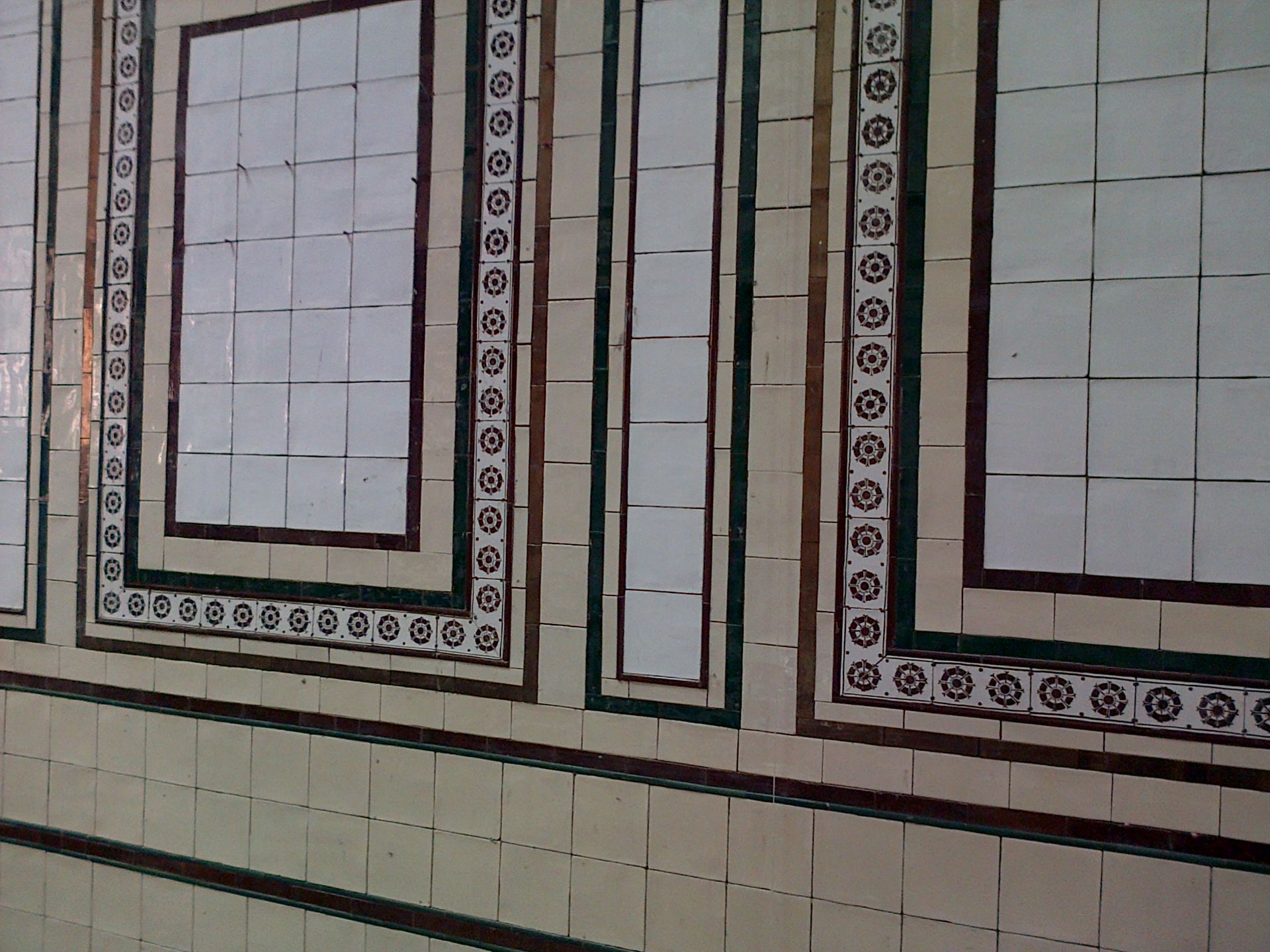 Pared de azulejos blancos con paneles enmarcados, bordes marrones y verdes, posiblemente el interior de un edificio.
