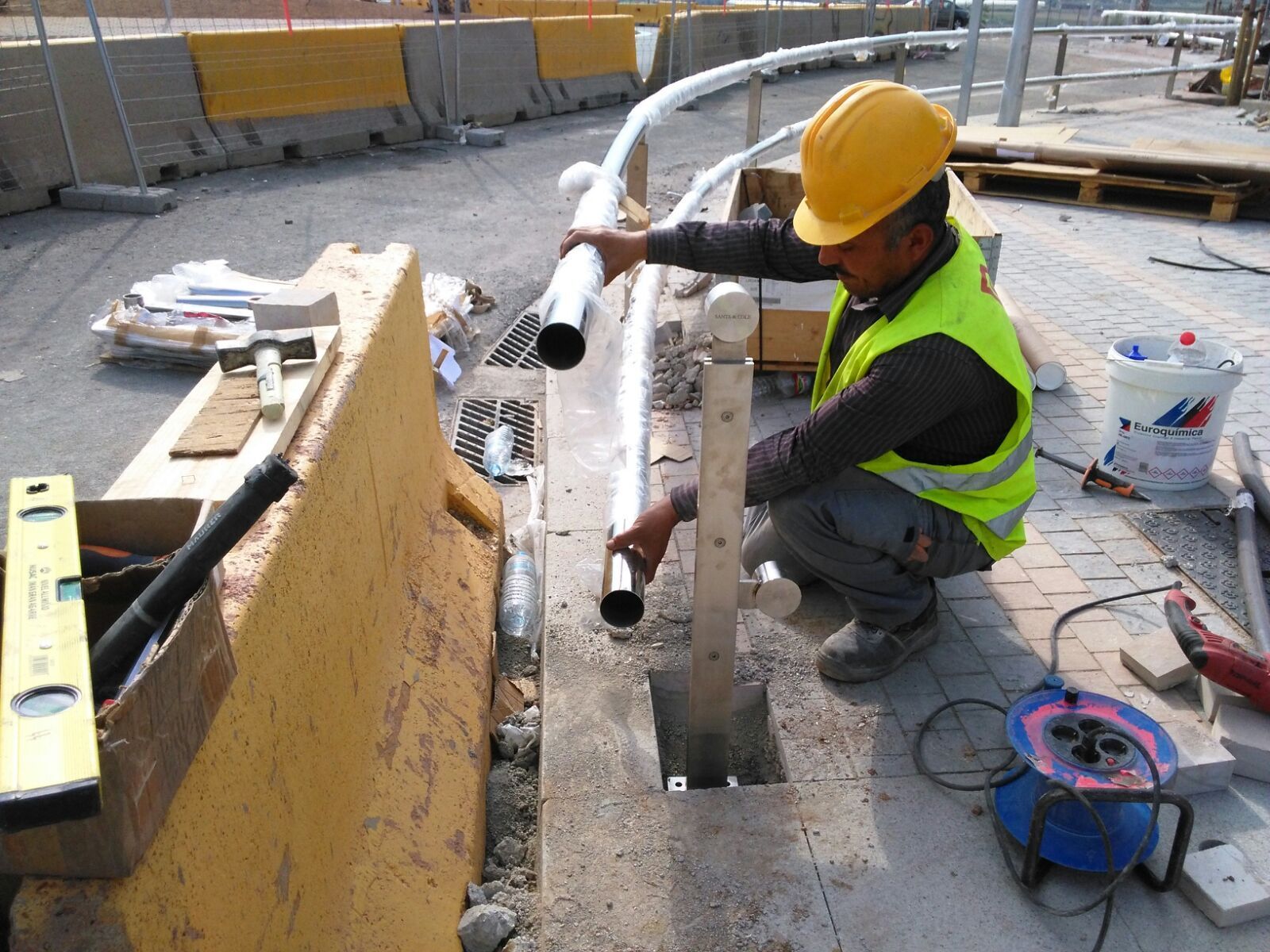 Un obrero instala una barandilla en una acera. Lleva casco y chaleco amarillos y se arrodilla.