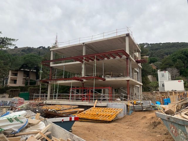 Edificio en construcción: estructura de hormigón de tres pisos con vigas de acero, emplazado sobre una ladera con árboles.