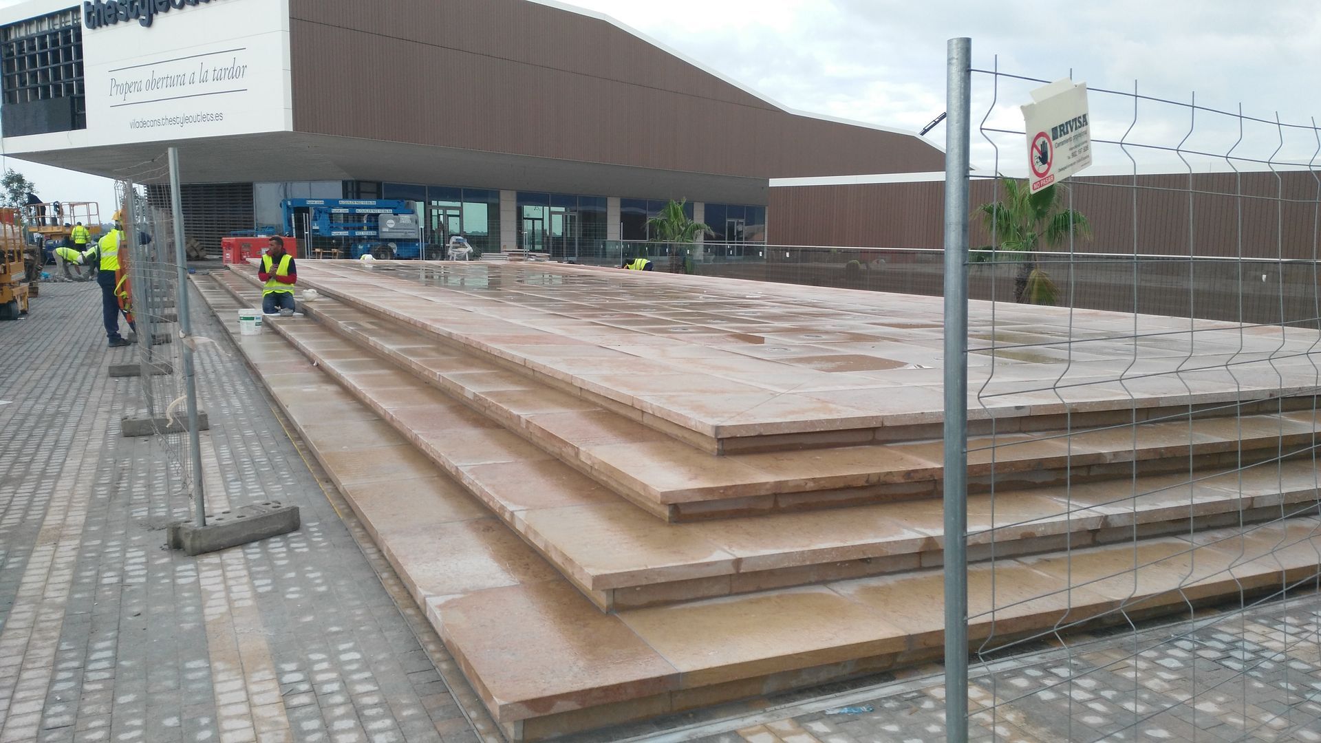 Obra con escalones de contrachapado frente a un edificio. Trabajadores con chalecos de seguridad. Valla alrededor de los escalones.