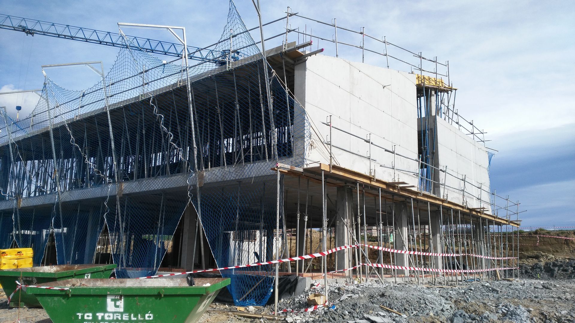 Edificio en construcción, rodeado de andamios y una grúa. Estructura de hormigón gris con un contenedor verde.