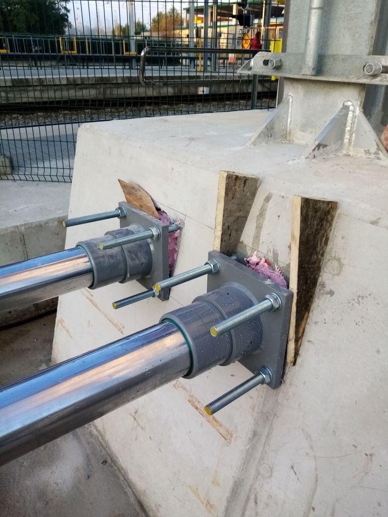 Pilar de hormigón con postes y soportes metálicos, fijado con pernos y cuñas de madera. Obra en el exterior.