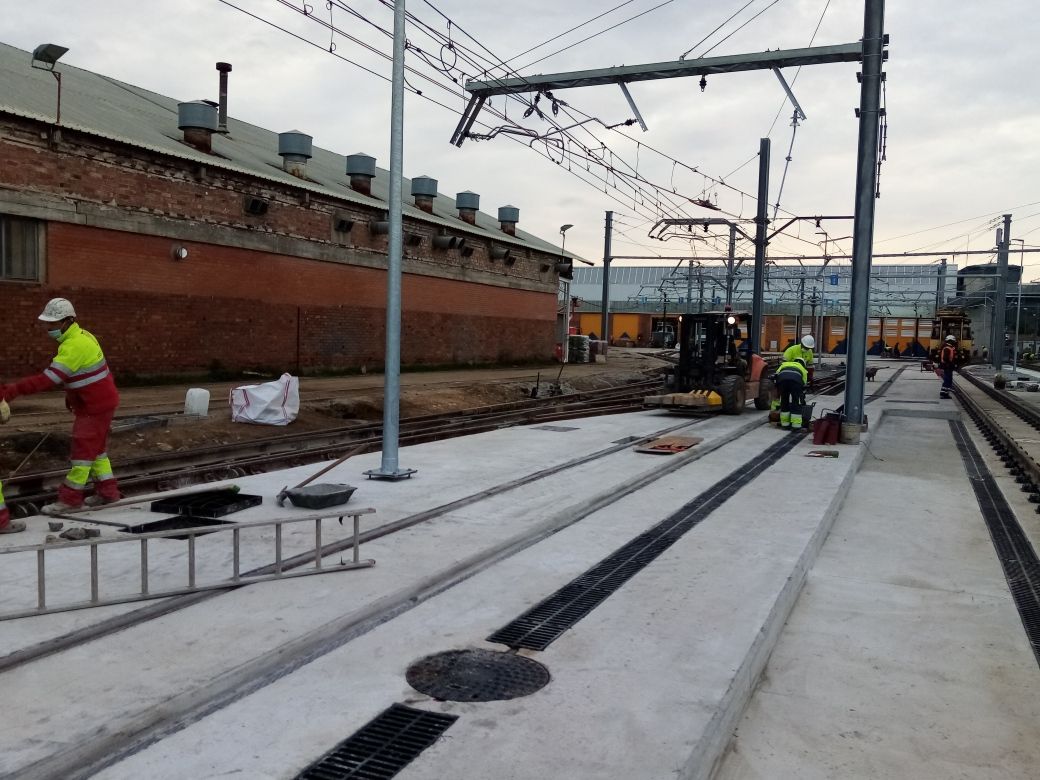 Trabajadores con equipo de seguridad en un andén ferroviario, cerca de vías y líneas eléctricas aéreas, junto a un edificio de ladrillos.