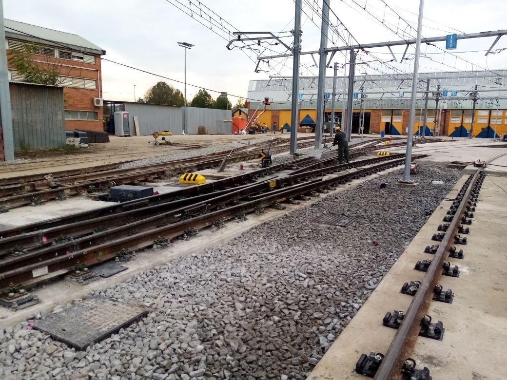 Vías ferroviarias con grava, líneas eléctricas aéreas y un edificio de ladrillo.