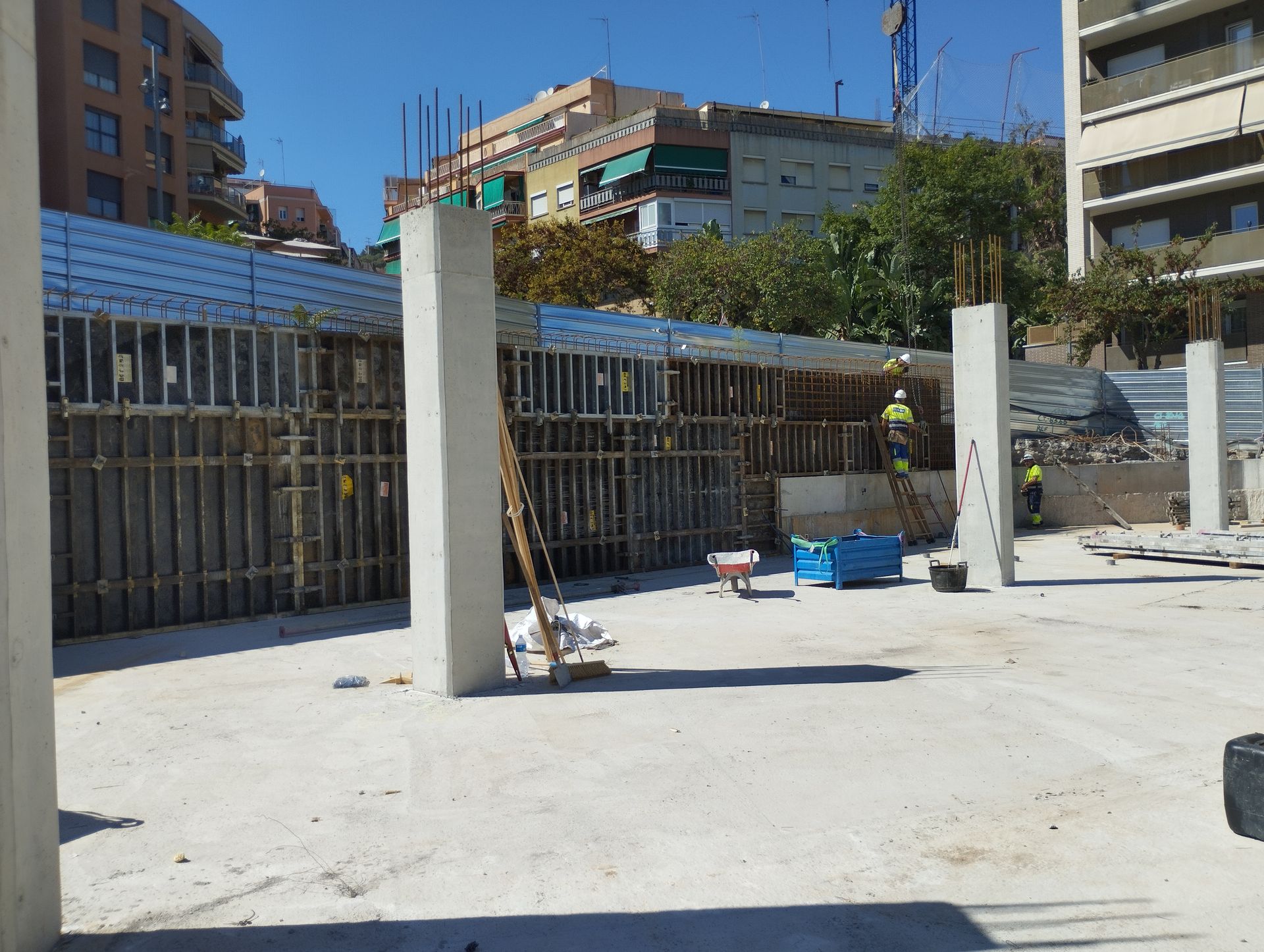 Obra con columnas y encofrados de hormigón. Trabajadores con chalecos amarillos. Edificios urbanos al fondo.