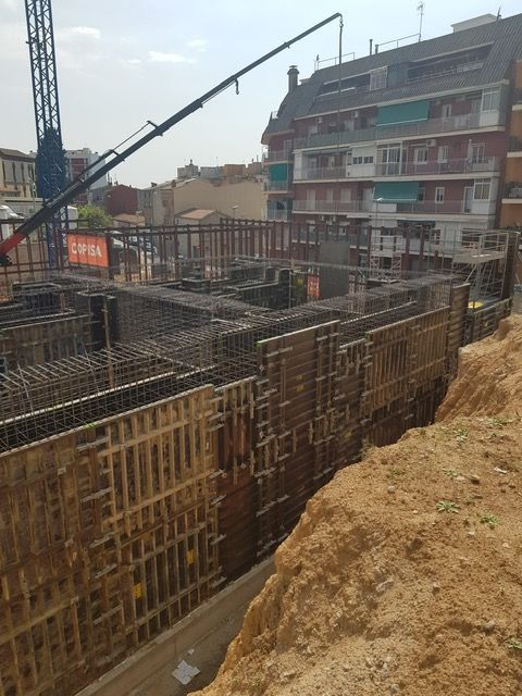 Sitio de construcción con encofrados de madera, refuerzo de acero, grúa, edificios al fondo.