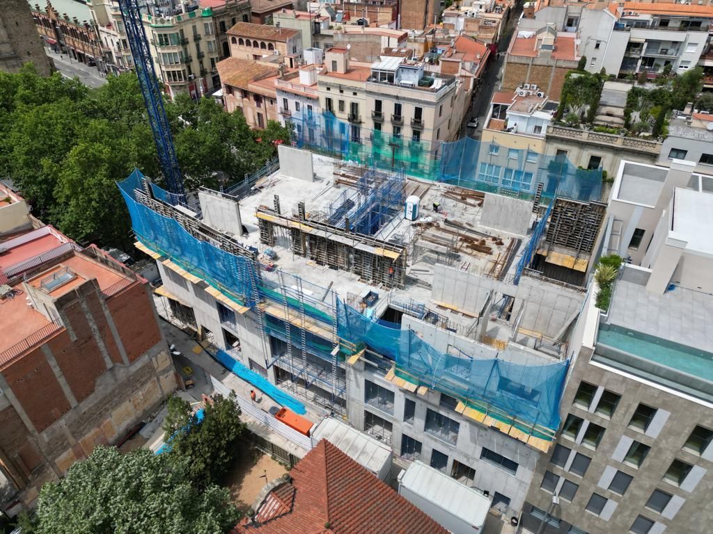 Vista aérea de un edificio en construcción en una ciudad, rodeado de otros edificios, con una grúa y red de seguridad.