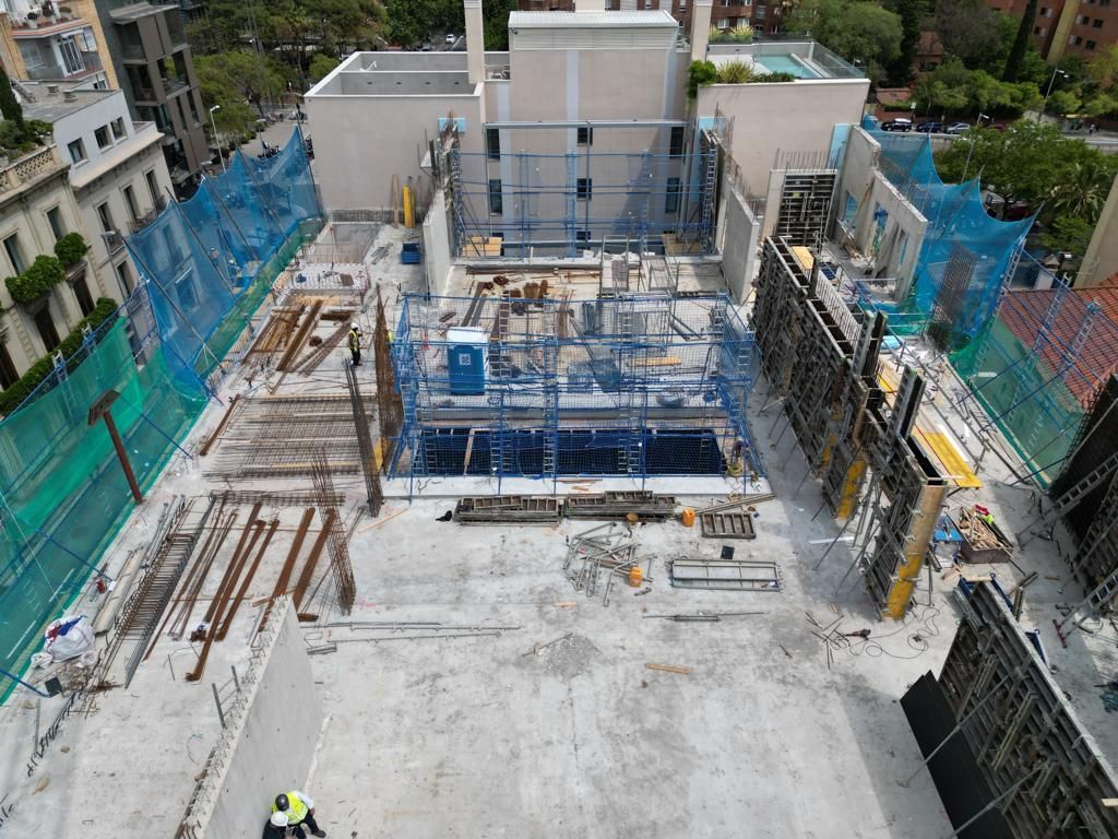 Sitio de construcción visto desde arriba, red de seguridad azul, hormigón, vigas de metal y un baño portátil.