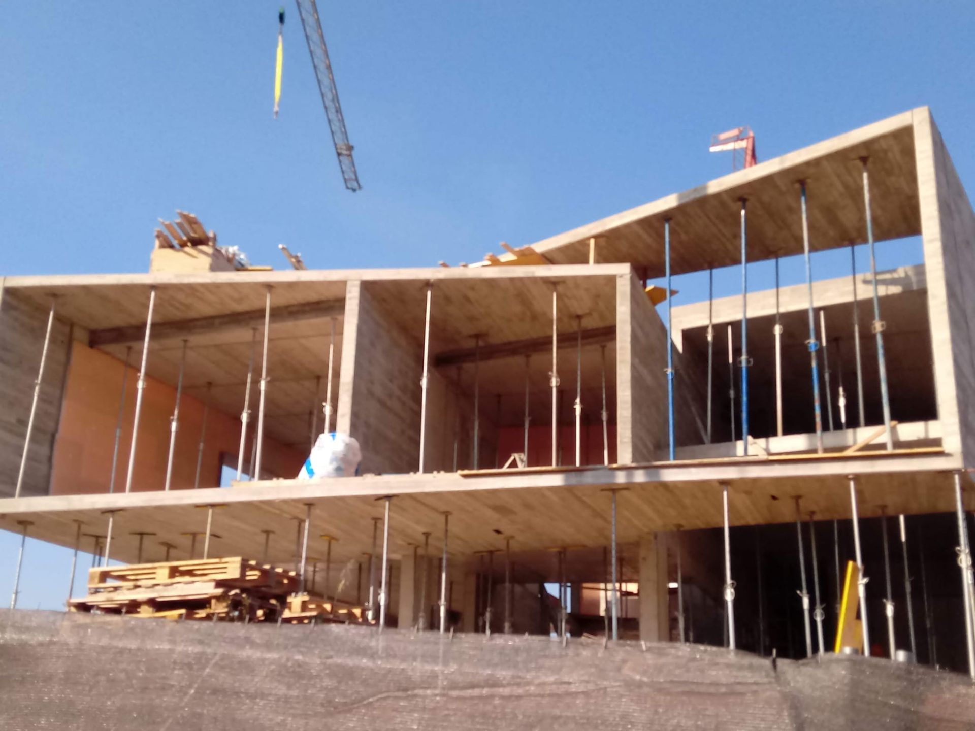 Edificio en construcción, estructura de hormigón sostenida por postes temporales, grúa al fondo.