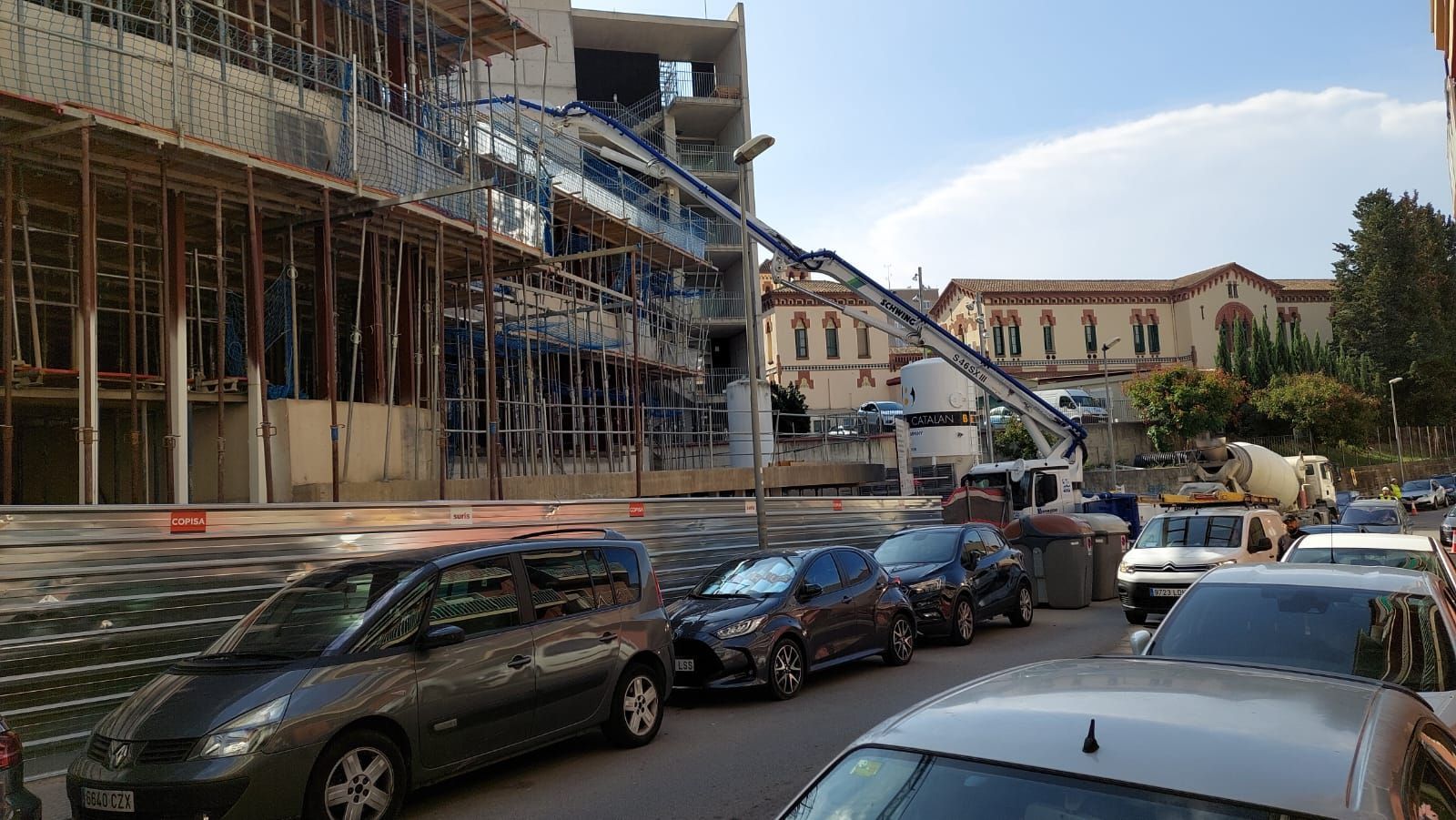 Sitio de construcción con el brazo de la bomba de hormigón llegando al edificio; autos estacionados a lo largo de la calle.