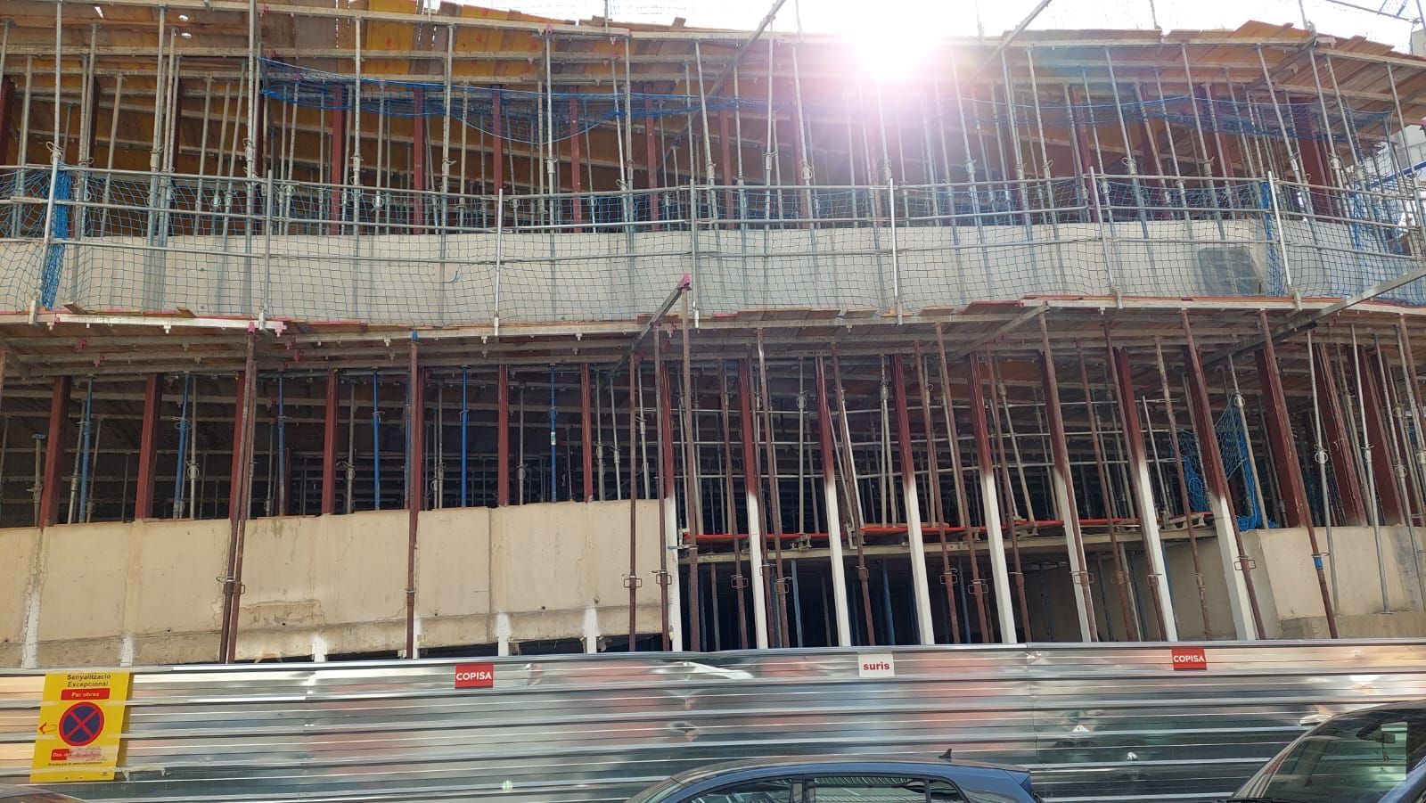 Obra con andamios y muros de hormigón. Vigas de soporte rojas y azules. Luz solar intensa.