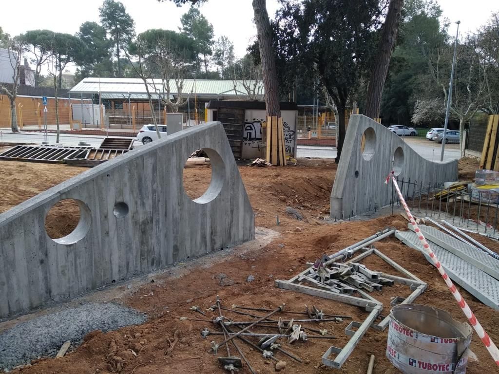 Entrada de parque de hormigón con recortes circulares en construcción; árboles y edificios al fondo.