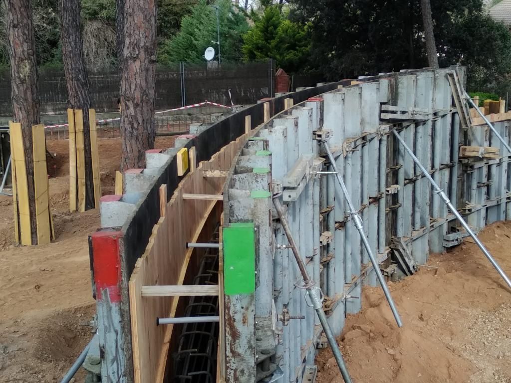 Sitio de construcción: muro de hormigón curvo con encofrados colocados, barras de refuerzo expuestas y estructuras de soporte.
