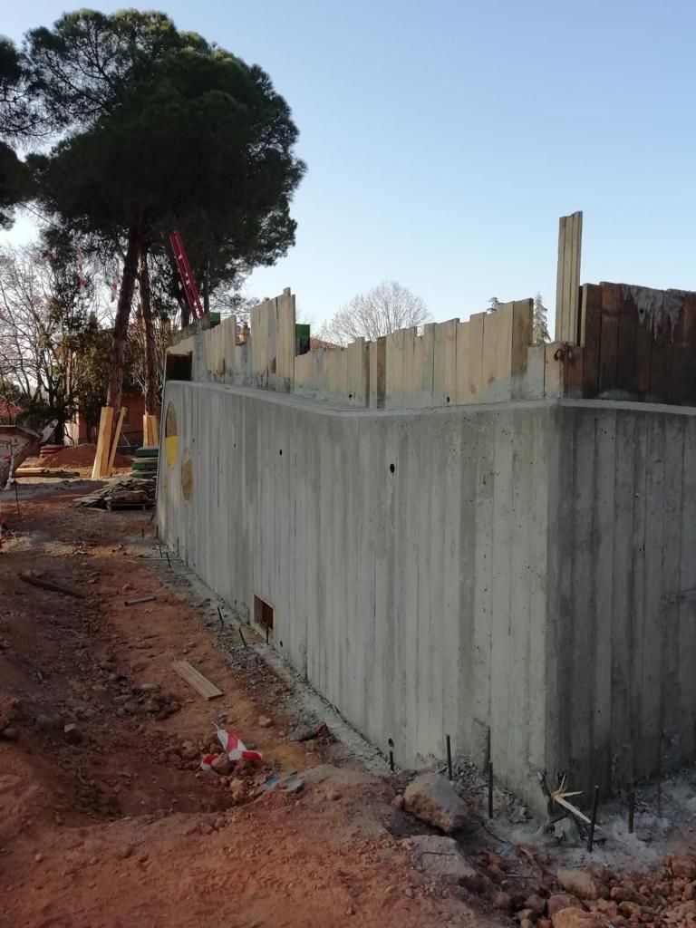 Muros de cimentación de hormigón de un edificio en construcción; suelo de tierra; árboles y cielo al fondo.