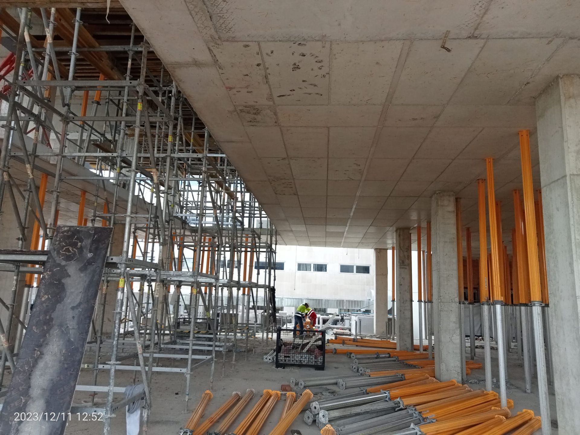 Interior de una obra con andamios, soportes y estructuras de hormigón sin terminar. Hay trabajadores presentes.