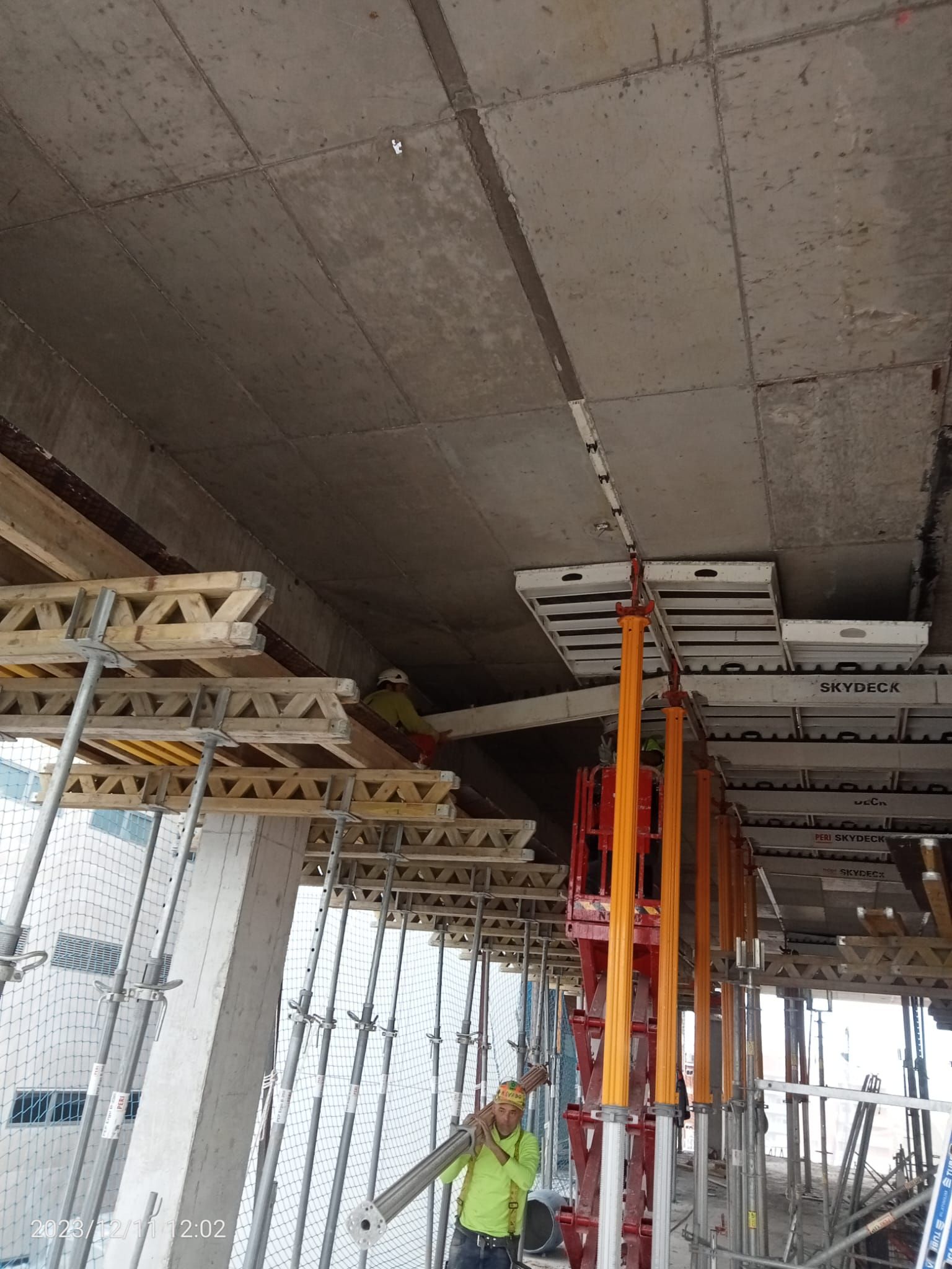 Obra: Obreros con herramientas bajo un techo de hormigón, sostenidos por andamios y correas.