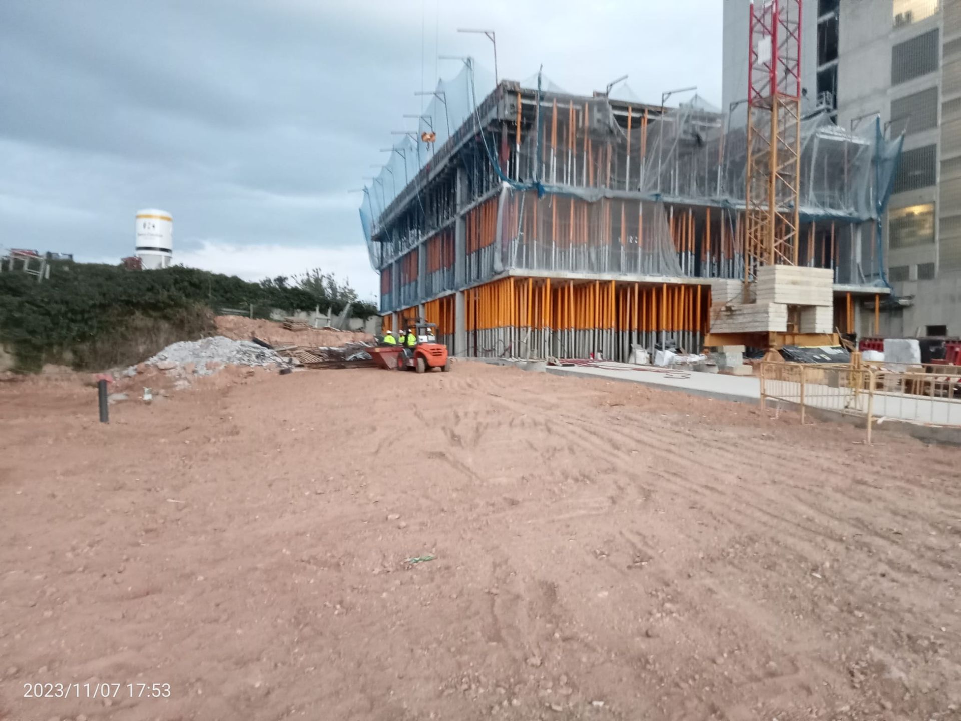 Obra con un edificio parcialmente construido, con soportes naranjas y red de seguridad. El suelo es de tierra.