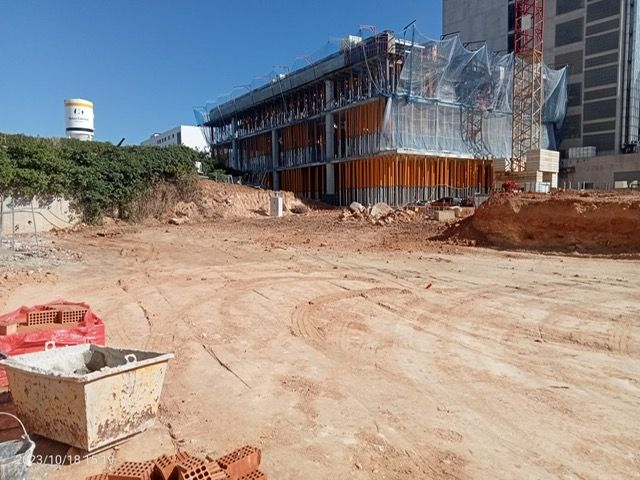 Sitio de construcción con edificio en desarrollo; tierra en primer plano, andamios, cielo azul.