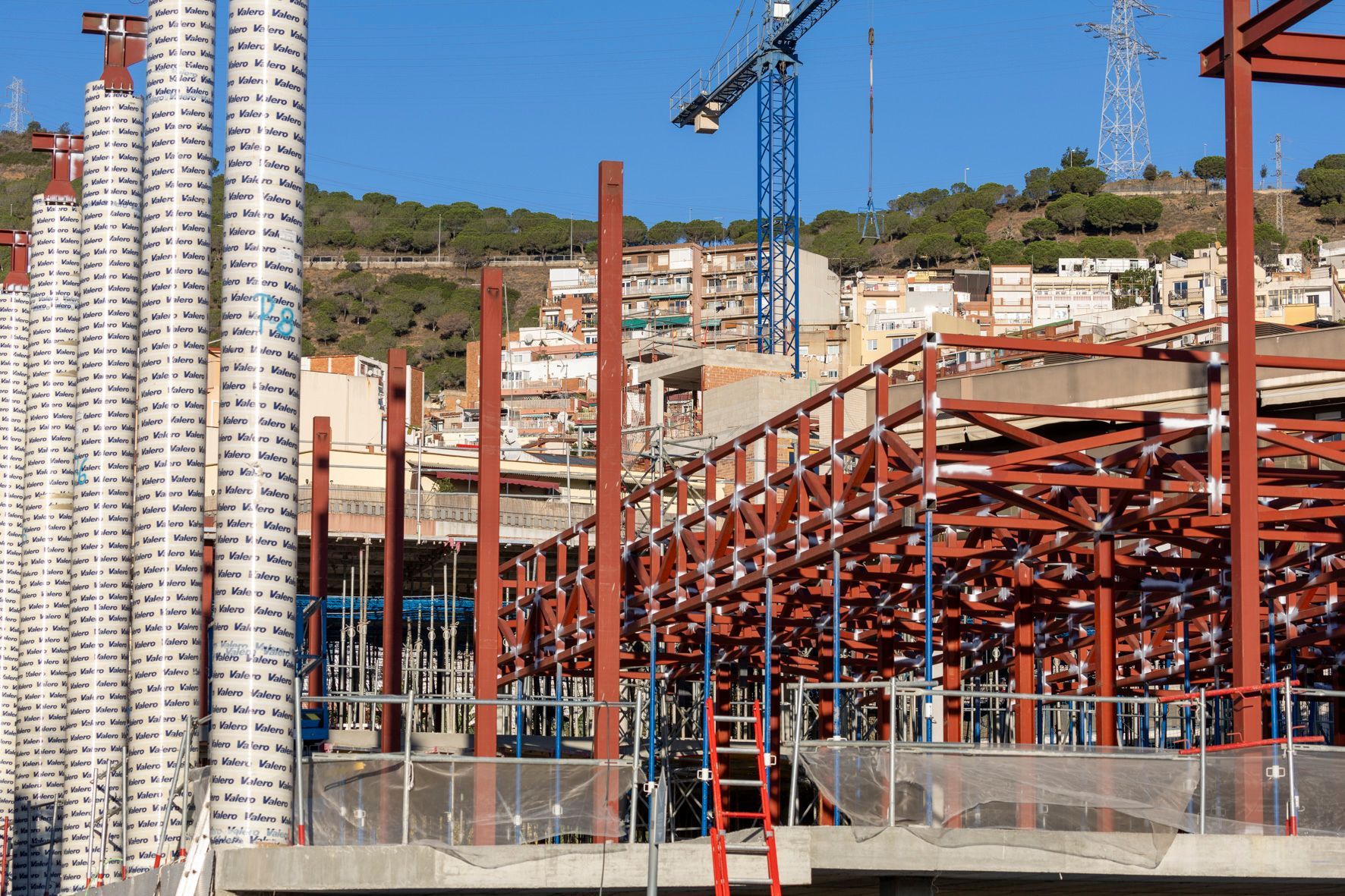 Sitio de construcción con estructura de acero roja, altas columnas blancas, grúa azul y edificios en la ladera al fondo.