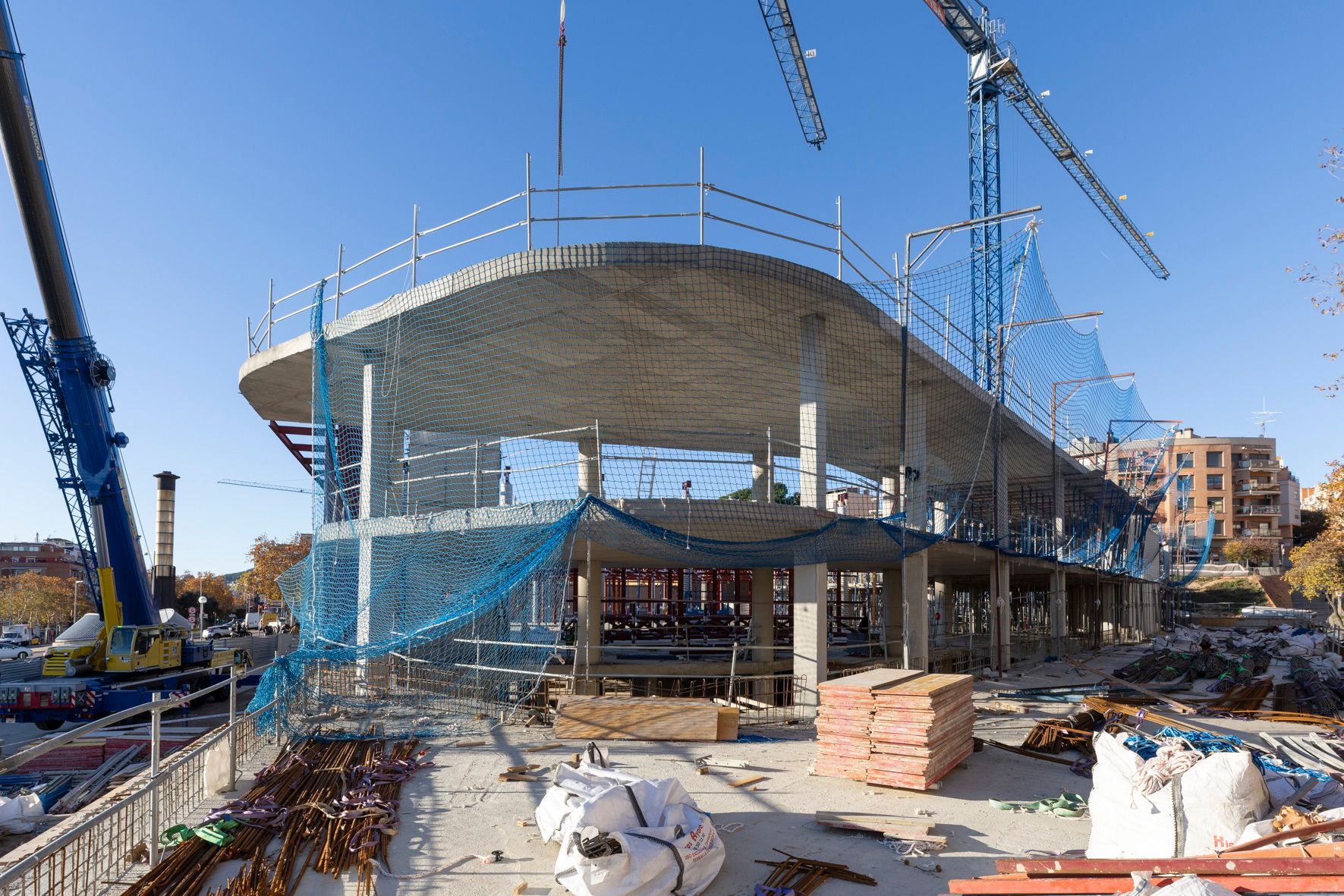 Obra de construcción de un edificio con techo curvo, rodeado de grúas y redes de seguridad en un día soleado.