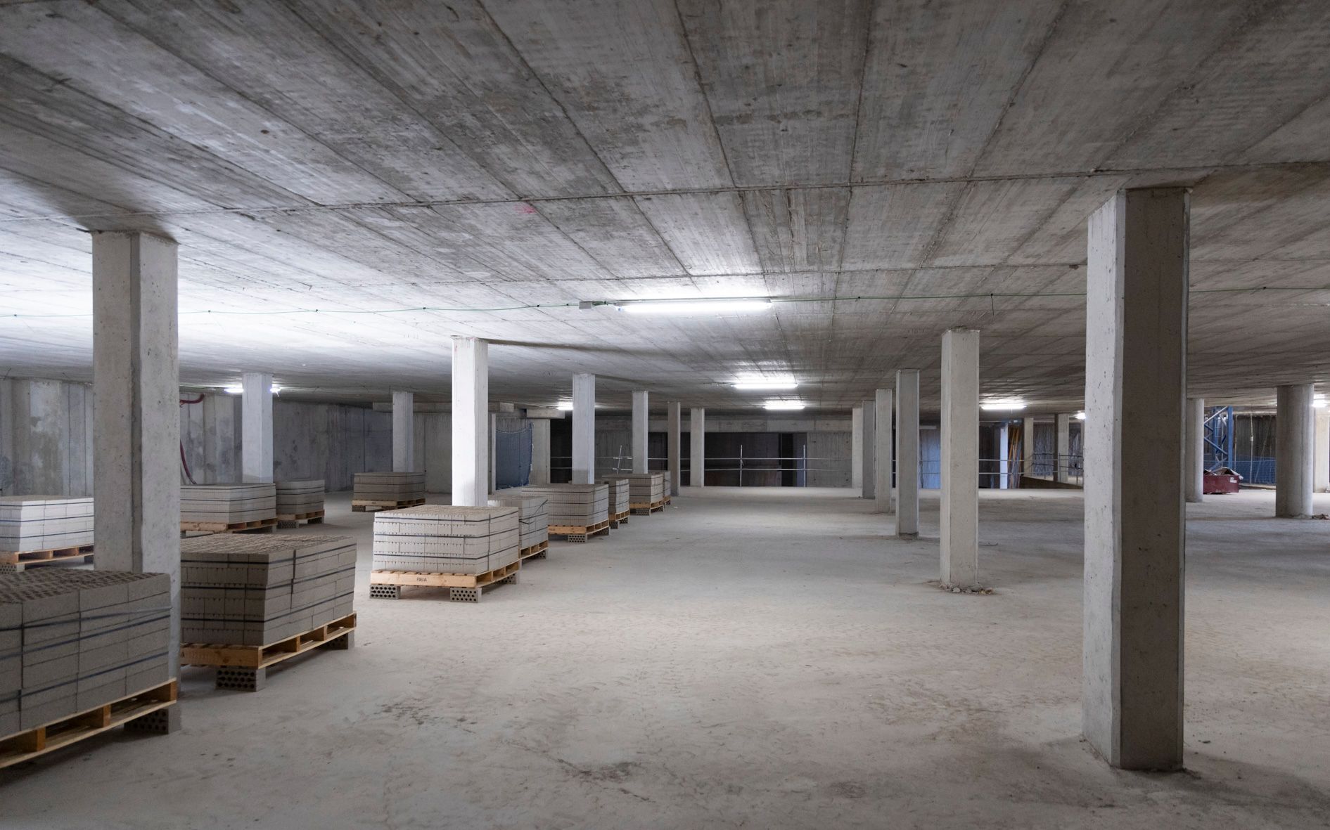 Obra interior de hormigón con pilares, palets de materiales e iluminación cenital.