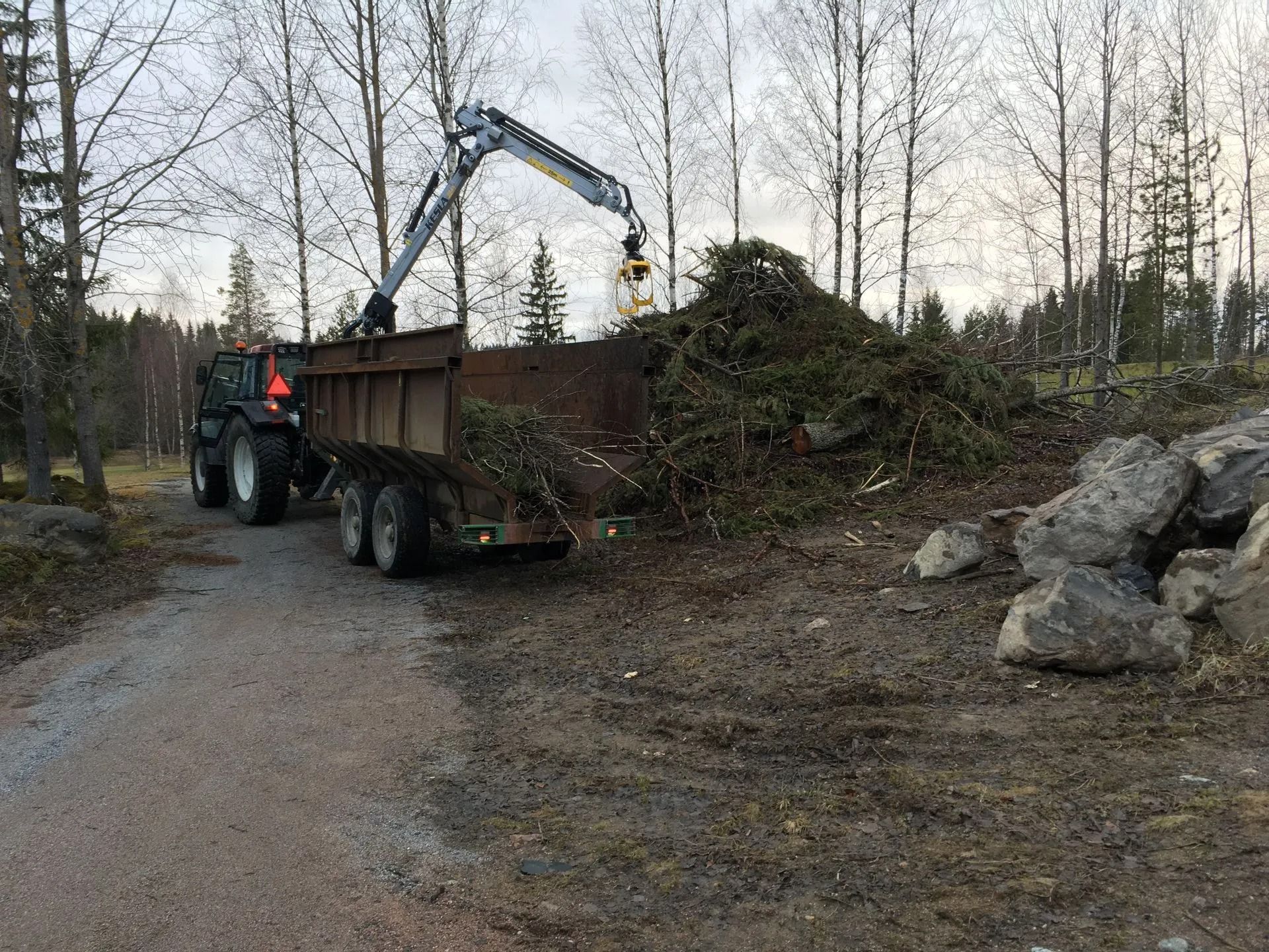 Traktori tukkiperävaunun ja nosturin kanssa lastaamassa harjaa soratien varrella metsäalueella