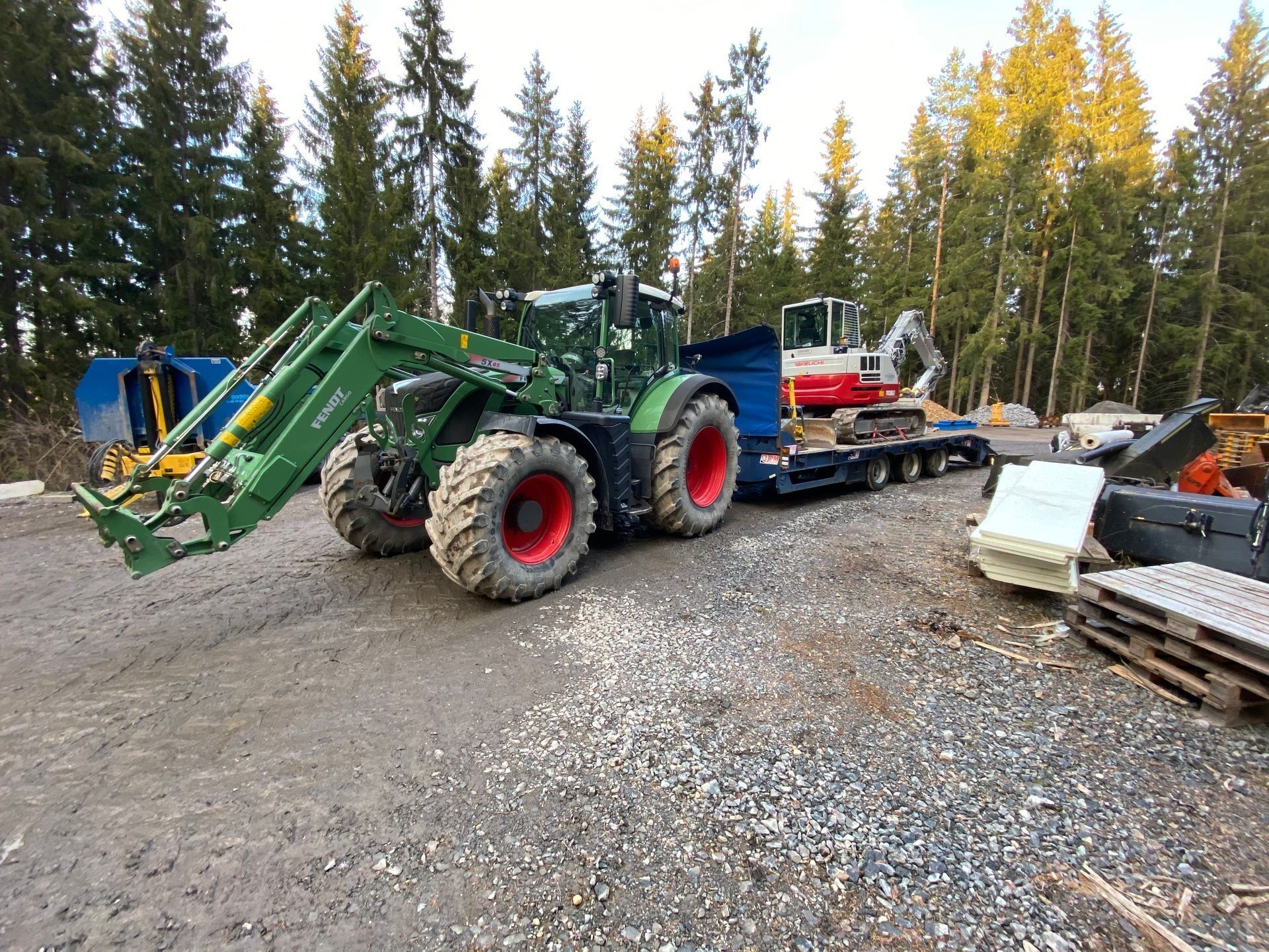 Vihreä traktori etukuormaajalla pysäköitynä sorakentälle laitteiden ja kuormalavojen viereen metsäalueella