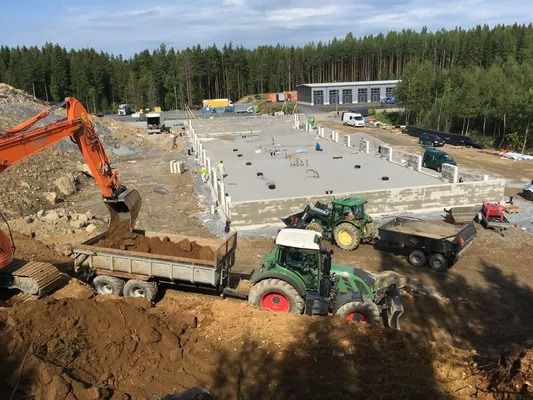 Rakennustyömaa, jossa kaivinkone ja vihreä traktori siirtävät maata betonialustan vieressä.